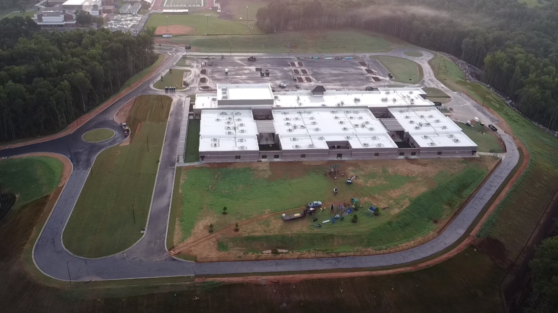 Aerial view of a large building under construction with surrounding roads, parking lot, and green spaces, some construction equipment and workers visible.