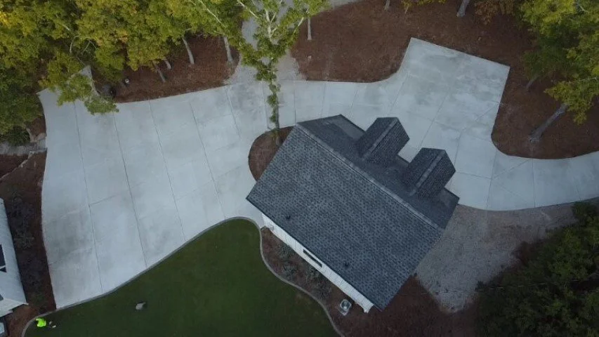 Aerial view of a modern house with a dark tiled roof, surrounded by a concrete pathway, trees, and a lawn.