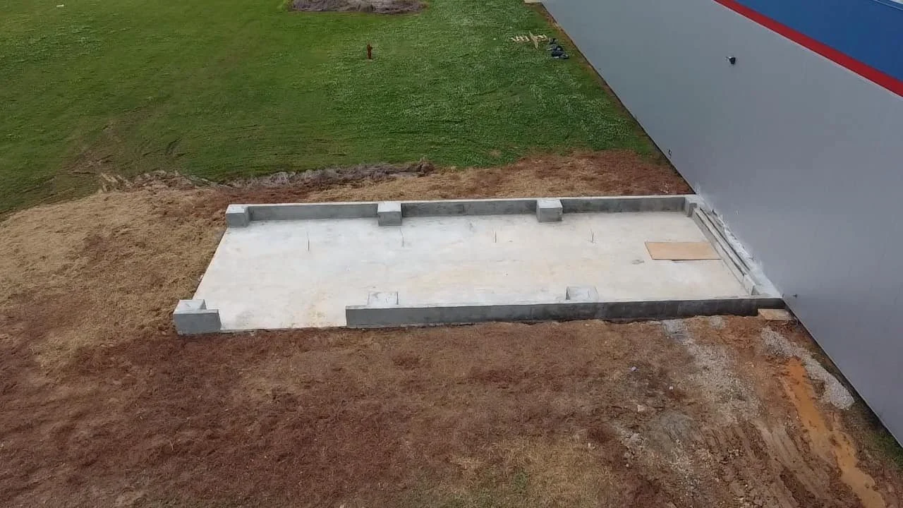 A construction site with a concrete foundation, surrounded by soil, next to a gray and blue building wall, and a grassy area in the background.