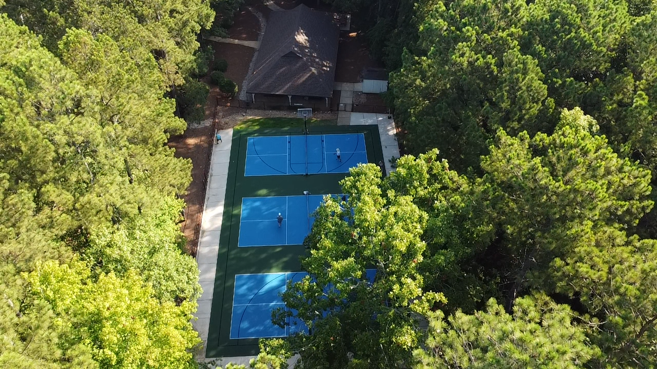 An aerial view of a blue outdoor basketball court surrounded by trees, with a building nearby, and a person seen on the court.
