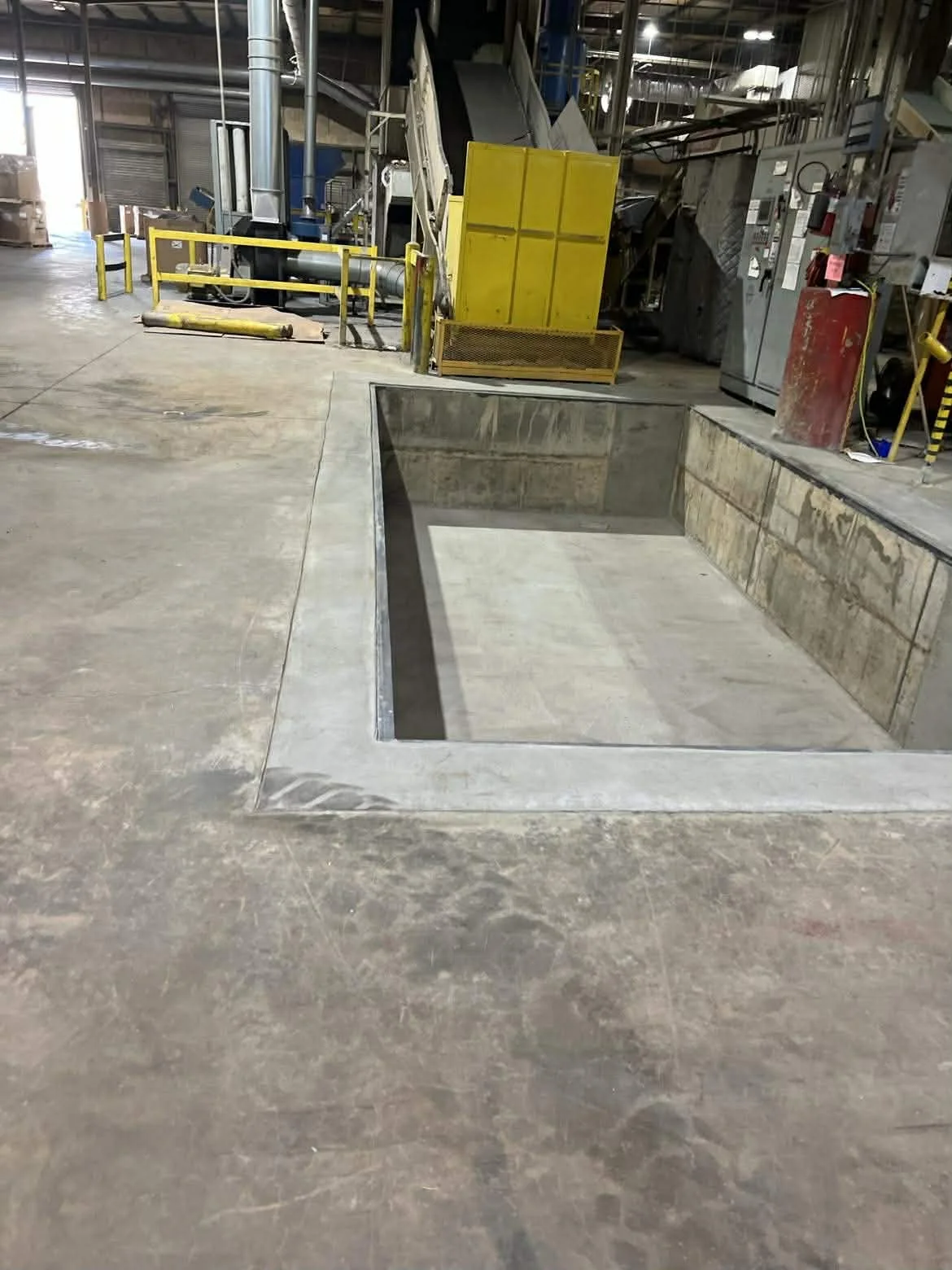 An industrial indoor space with a large rectangular empty concrete trench or pit in the floor, surrounded by construction or maintenance equipment and machinery.