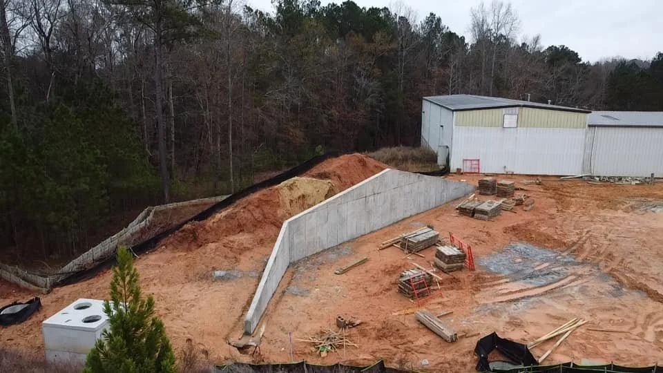 Construction site with a small retaining wall, dirt mounds, and a silver building in the background.