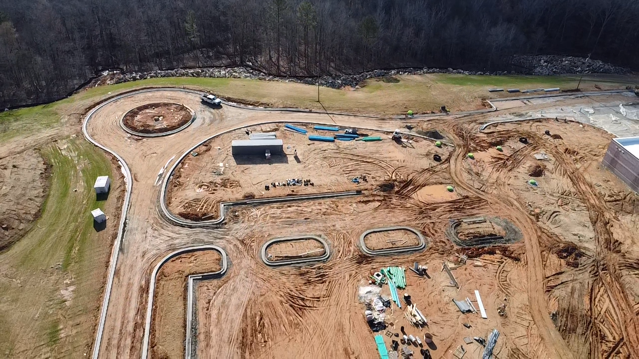 Aerial view of a construction site with winding paved paths, construction materials, and vehicles, surrounded by trees.