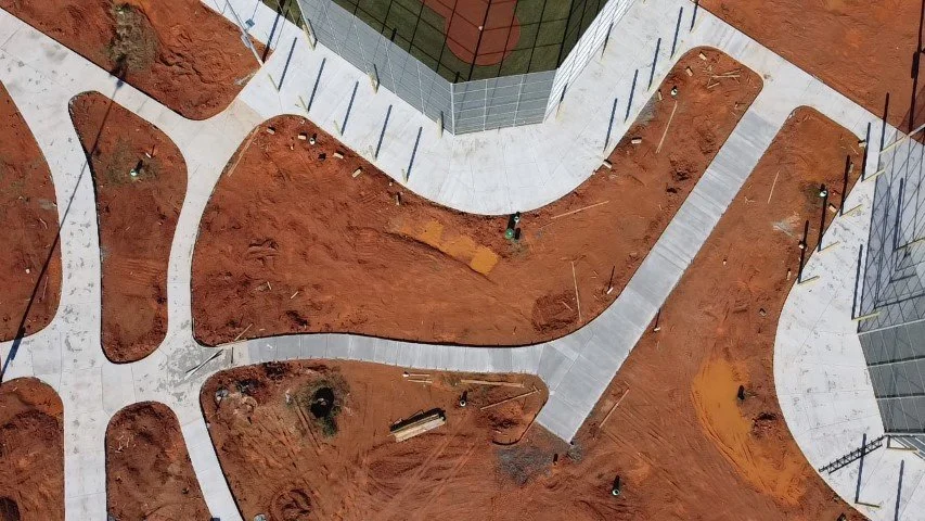 An aerial view of a construction site with newly poured concrete sidewalks, surrounding dirt areas, and glass buildings in the background.