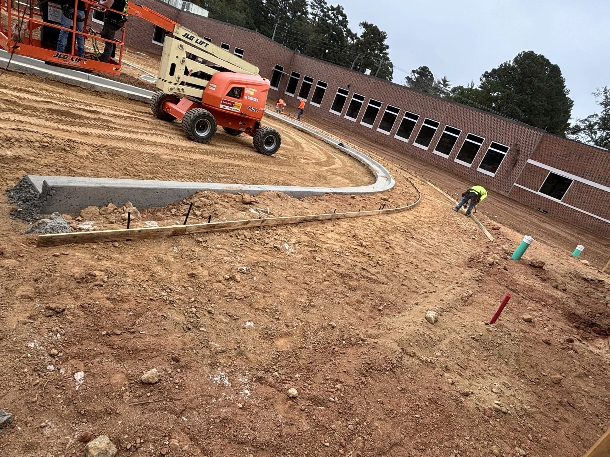 Construction site with workers and machinery preparing the area, with a brick building and trees in the background.