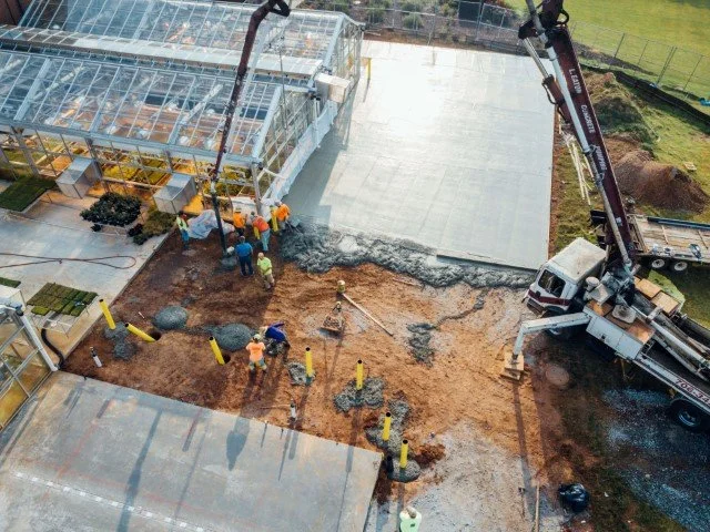 Construction workers laying concrete on a pathway at a construction site, with a concrete mixer truck pouring concrete and a glass greenhouse structure nearby.