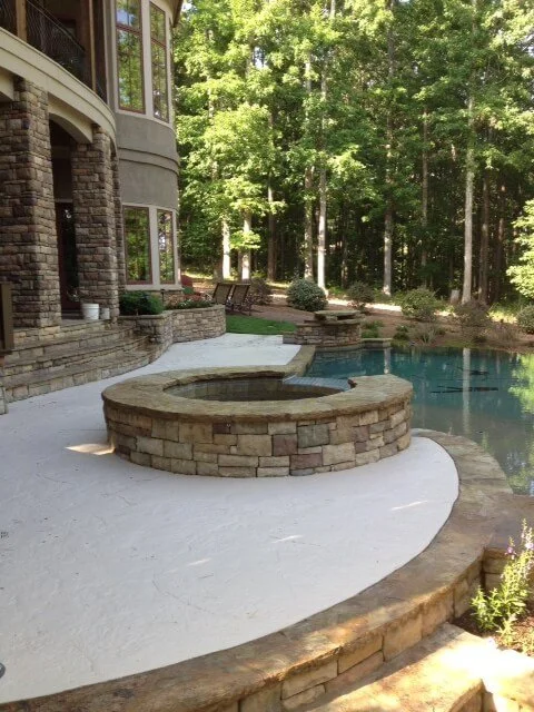 Backyard patio with a built-in hot tub, surrounded by stone and concrete, adjacent to a swimming pool, with a wooded backyard.