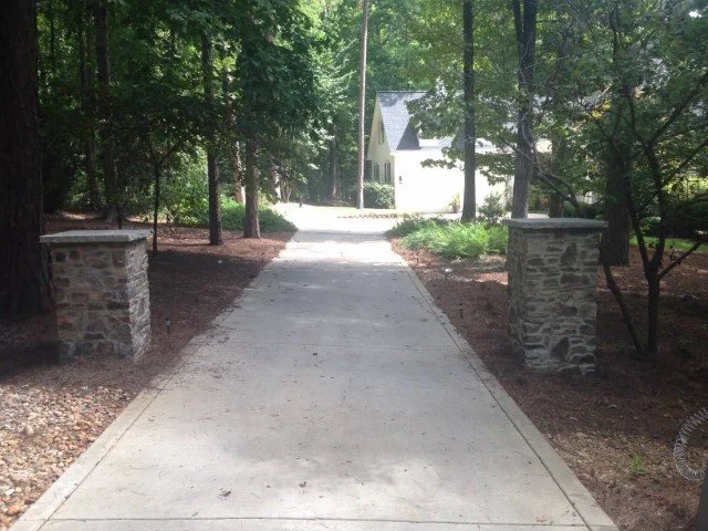 Concrete driveway leading to a house surrounded by trees, with stone pillars on either side at the entrance.