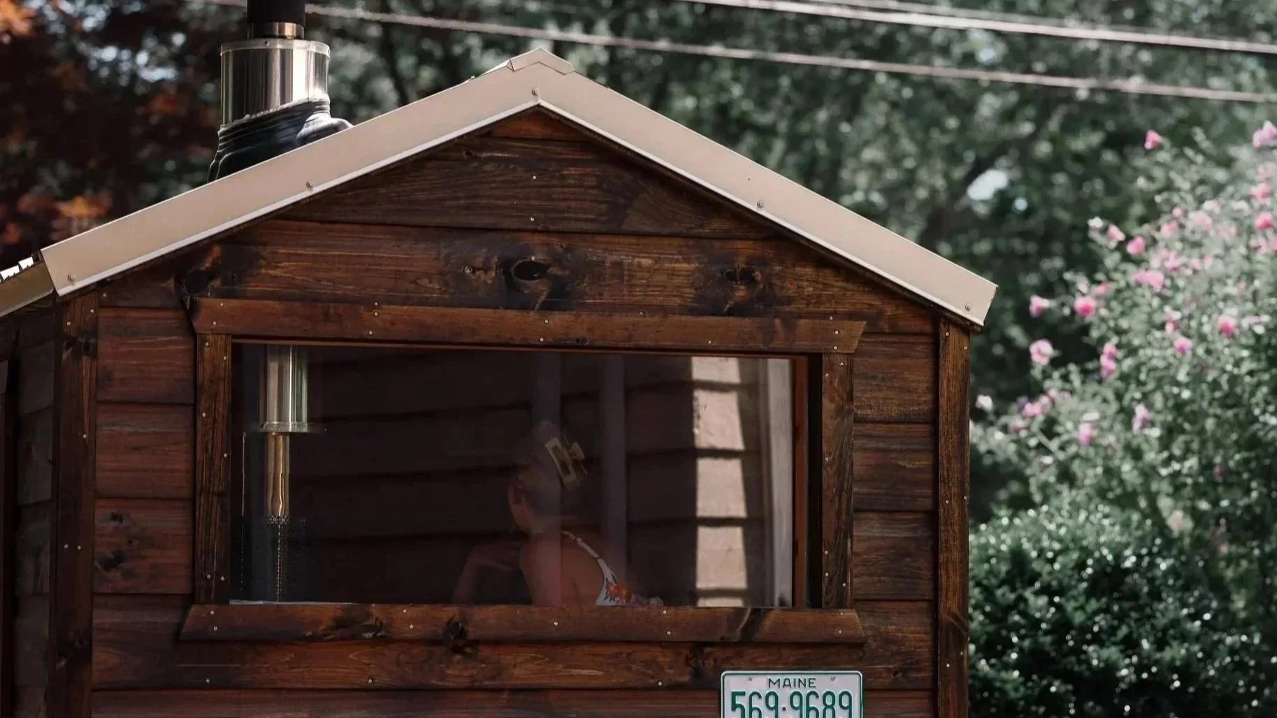 A small wooden house with a metal roof and a chimney, with a person visible through the window, and a Maine license plate reading 569-9689 at the bottom.