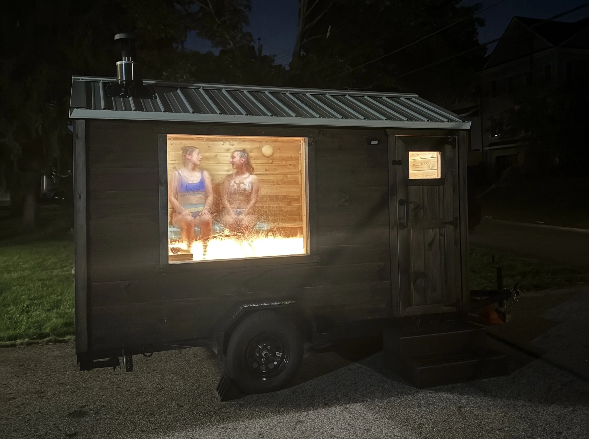 A brown outdoor wooden sauna with a metal roof and large glass window showing two women sitting inside, illuminated by warm interior lighting, at night. The women are smiling at each other and appear to be relaxing next to a fire or heater.