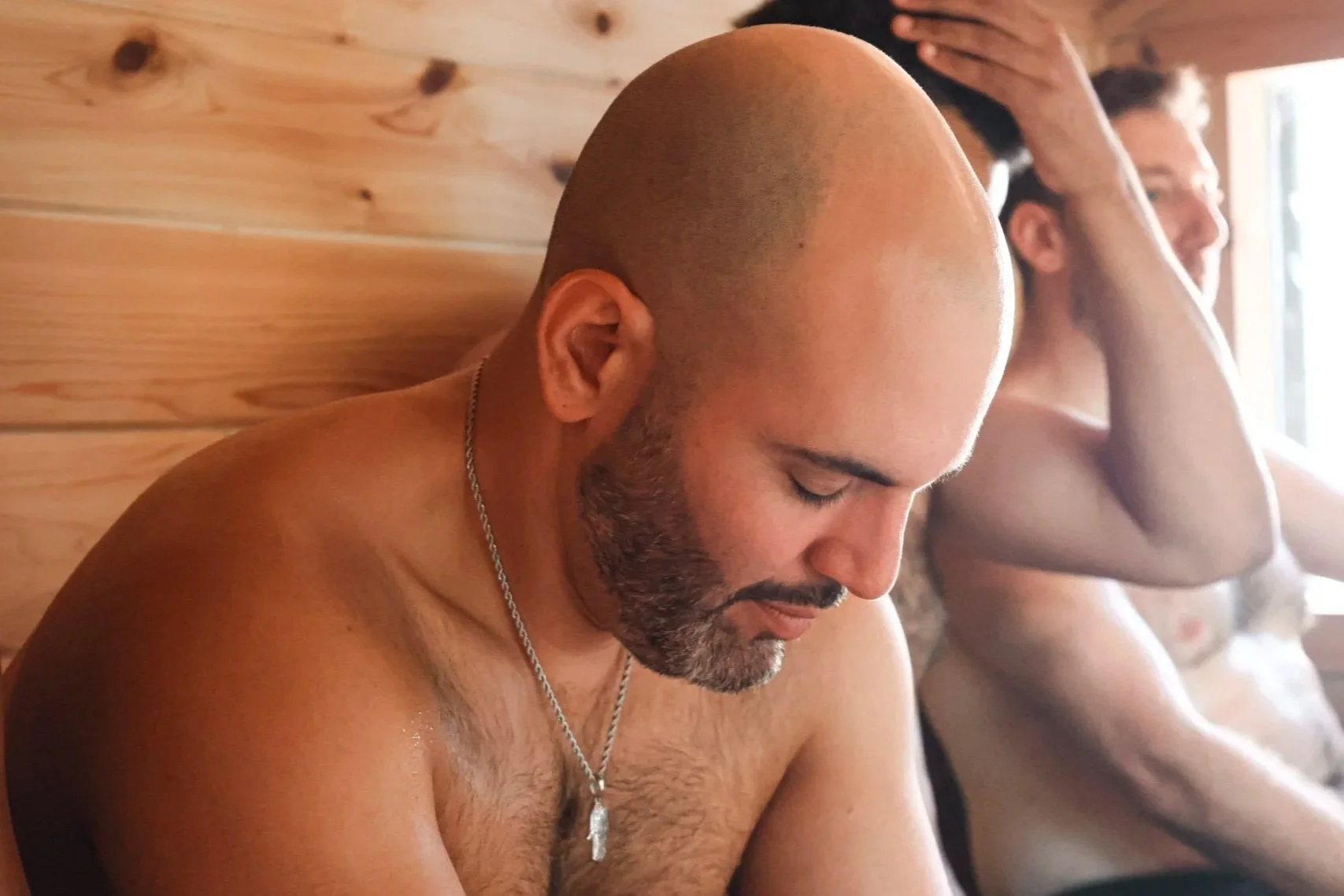 Two shirtless men sit with their eyes closed and hands on their heads in a wooden sauna.