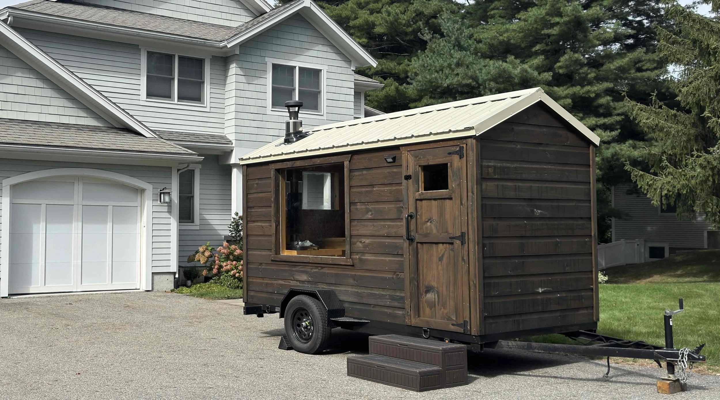 A sauna trailer with a window, parked in a residential driveway in front of a light gray house with white trim and a garage door. There are trees and bushes nearby.