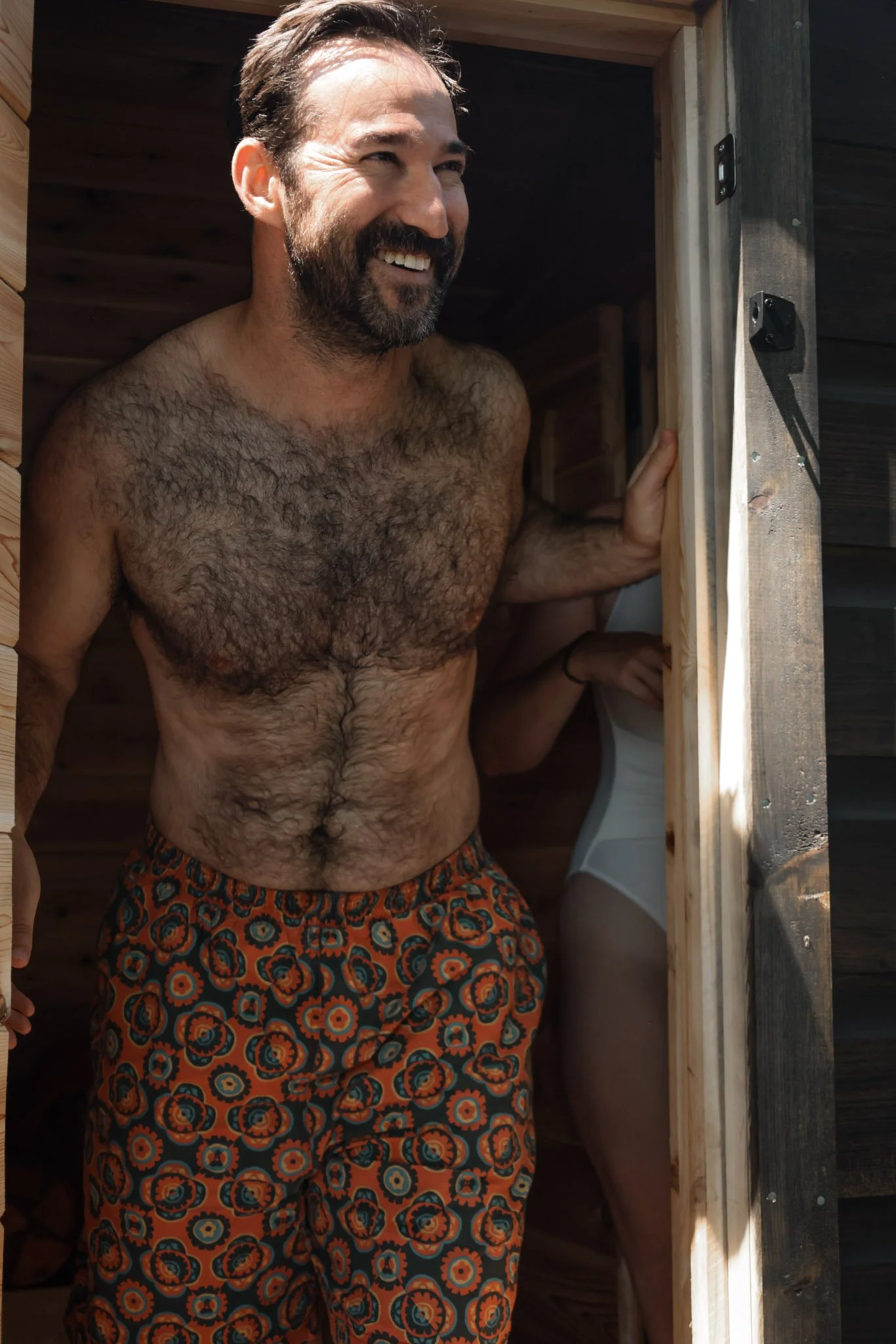A shirtless man with a beard and dark hair smiling while standing inside a wooden sauna, wearing colorful patterned shorts.
