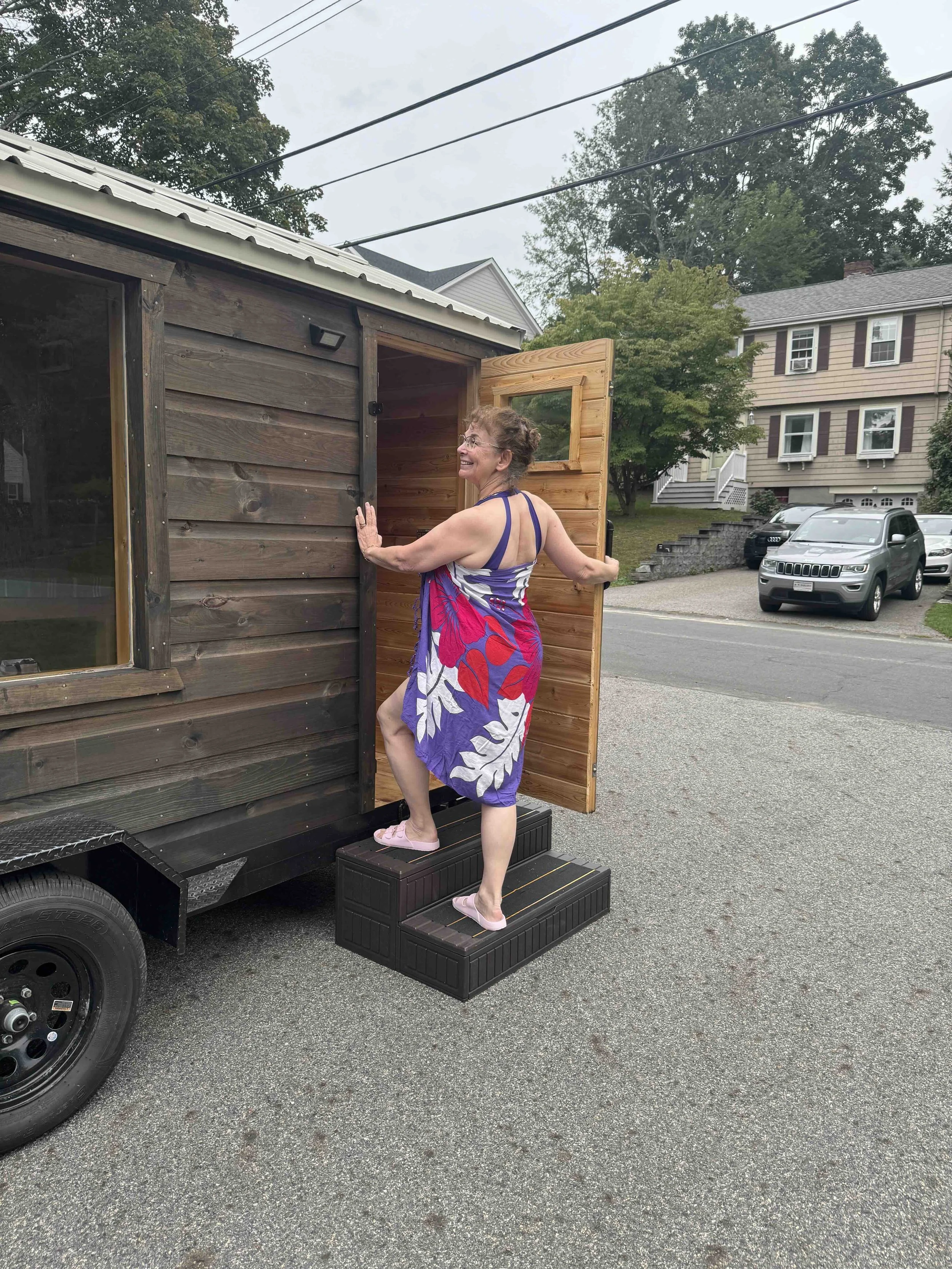A woman in a colorful dress is stepping onto a small stairway, entering a sauna trailer, smiling and holding the door open.
