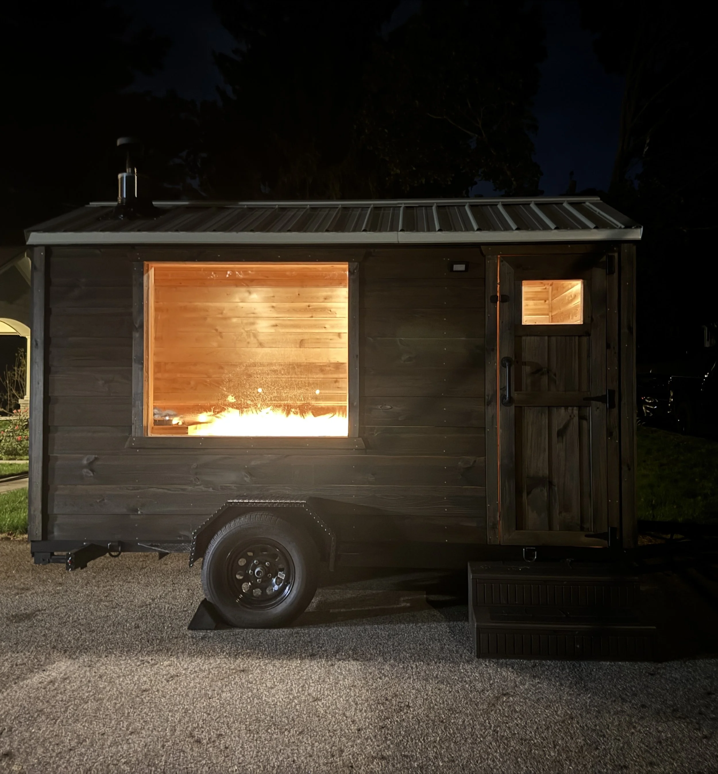 Nighttime photo of a sauna parked in a residential driveway, showing warm interior lighting, parked on a gravel surface with trees in the background.