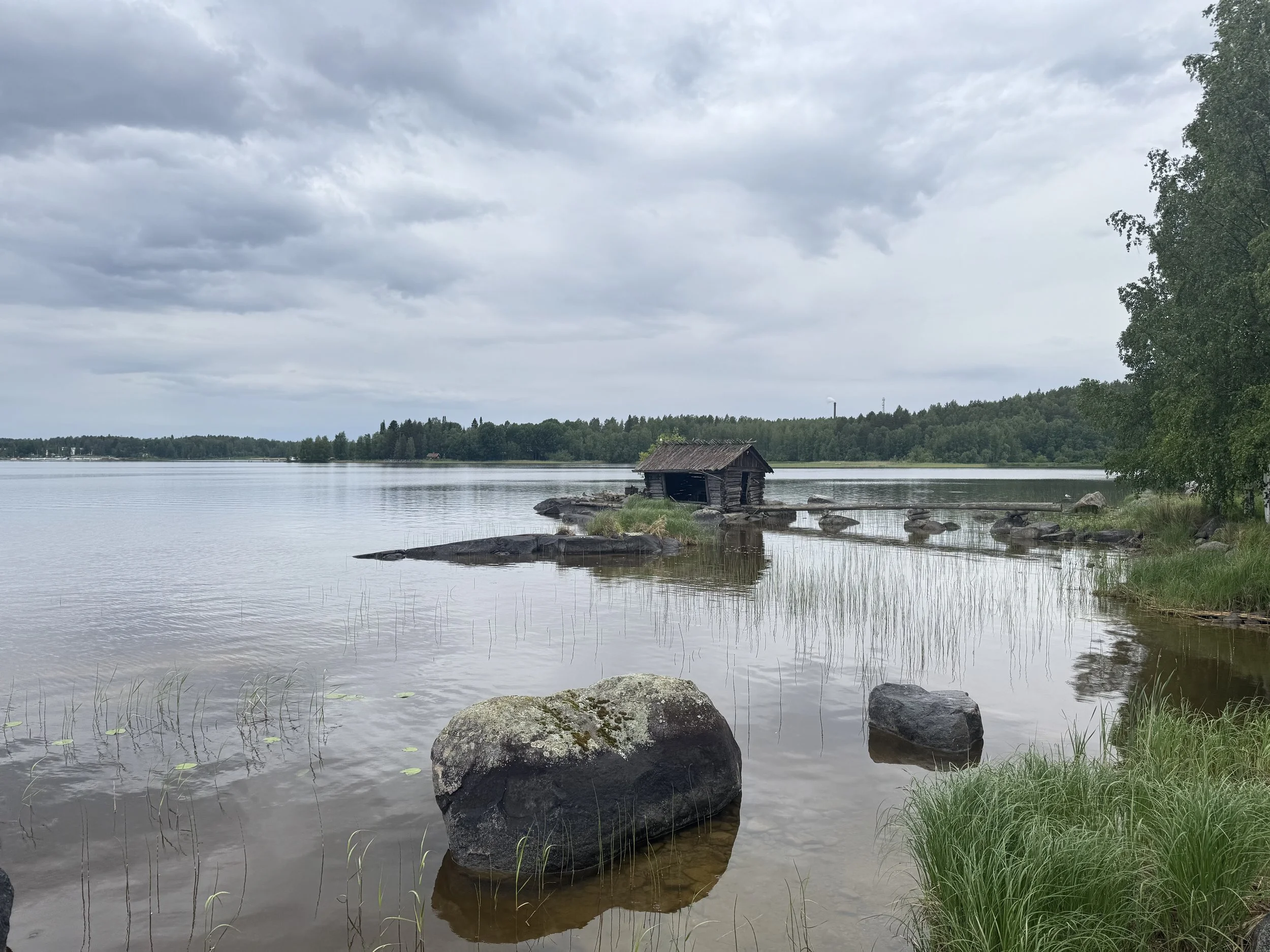 Overcast sky over a calm lake in Finland with a small, weathered wooden sauna structure on the shoreline, surrounded by water, rocks and grass, with a dense forest in the background.