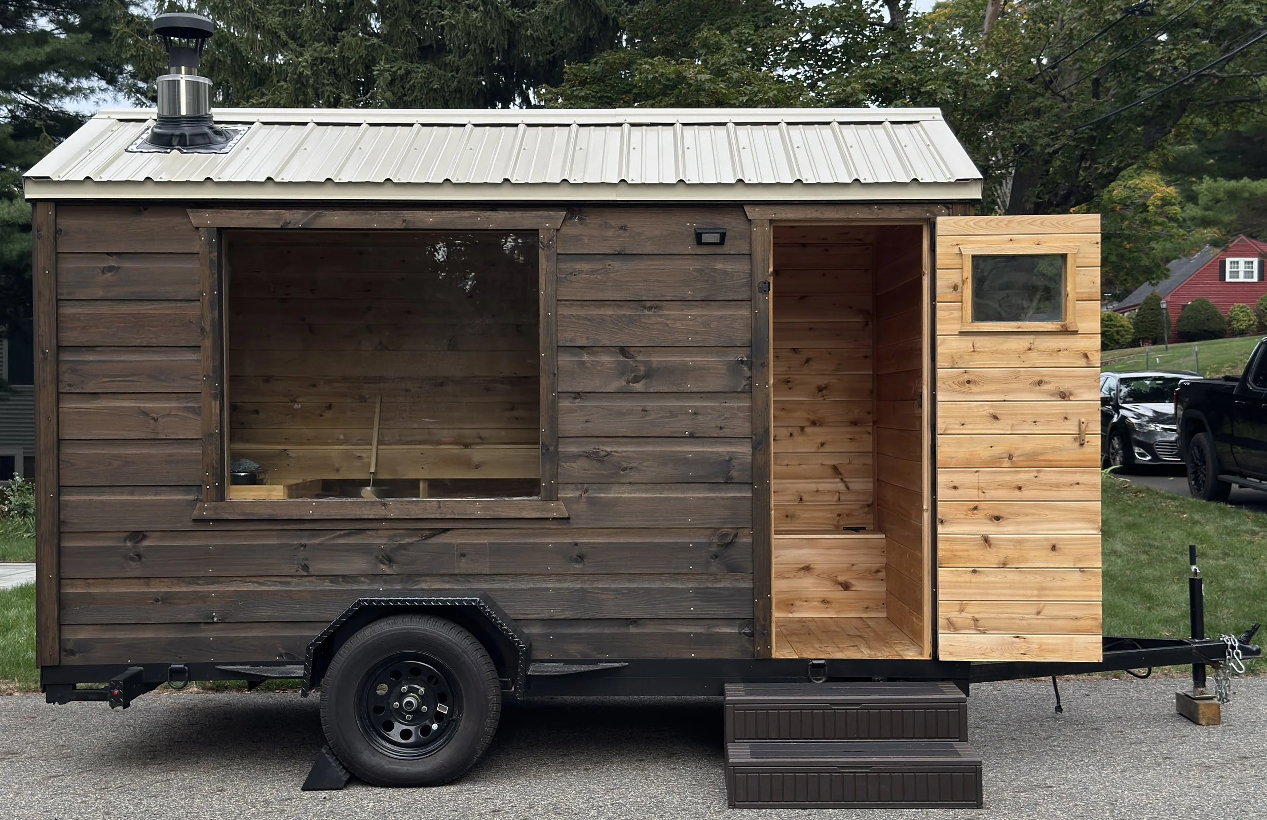 A sauna trailer with a window, and door is open, parked in a residential driveway. There are trees and bushes nearby.