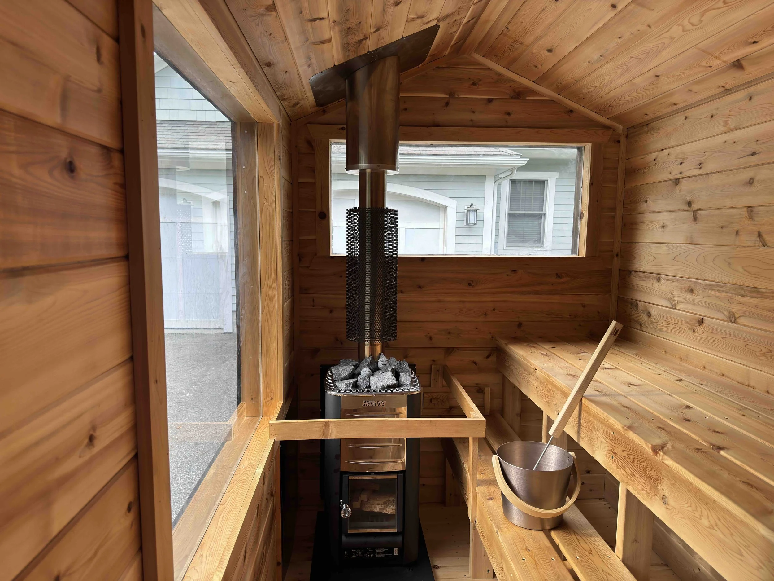 Interior of a small wooden sauna with windows, a smokestack, and a heating stove with rocks.