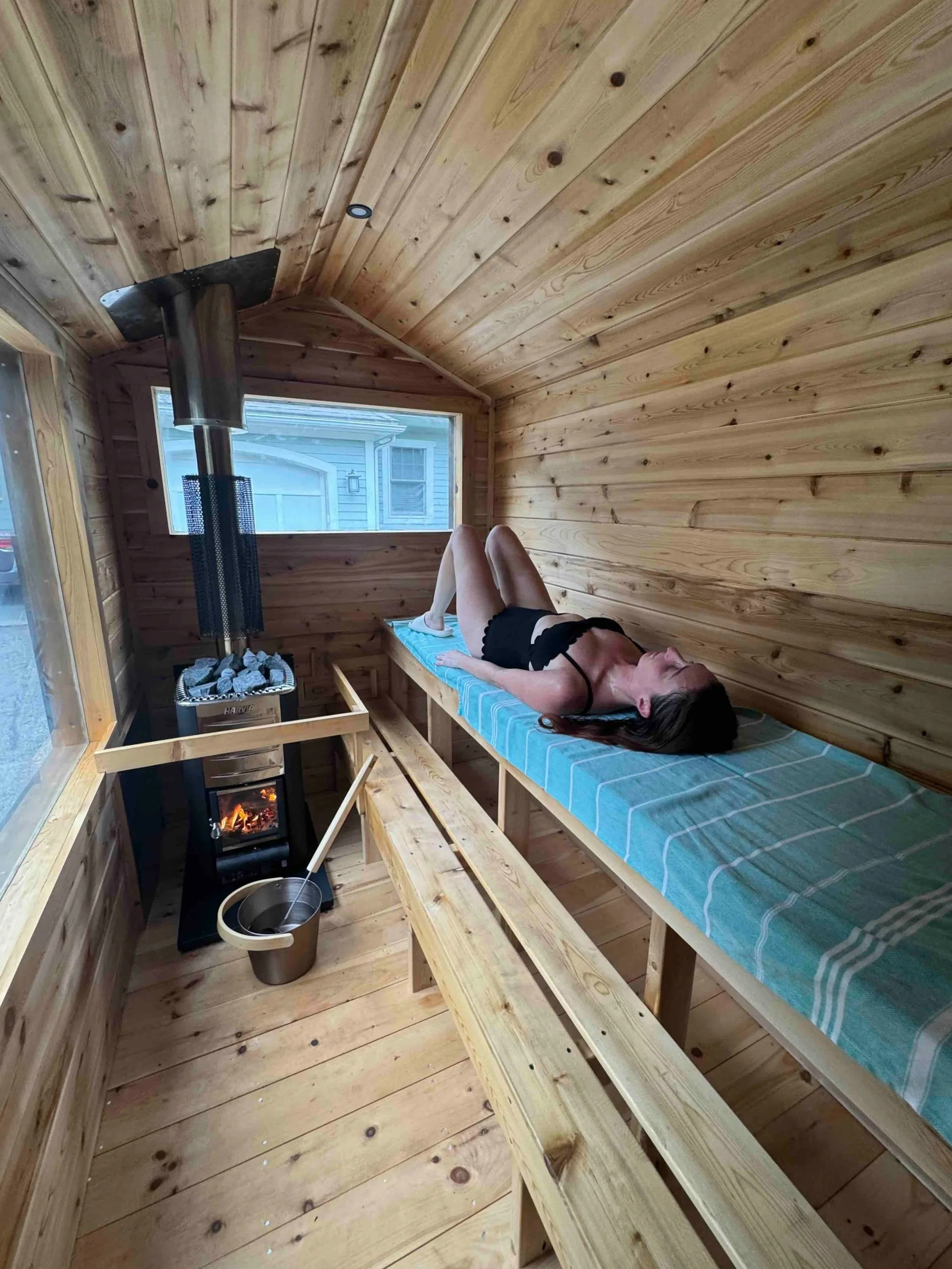 A woman lying on her side on a blue and white striped cushioned wooden bench inside a small wooden sauna. The room has wood-paneled walls and ceiling, a small window showing the outdoors, a stove with rocks and a chimney, and a small bucket with a ladle nearby.
