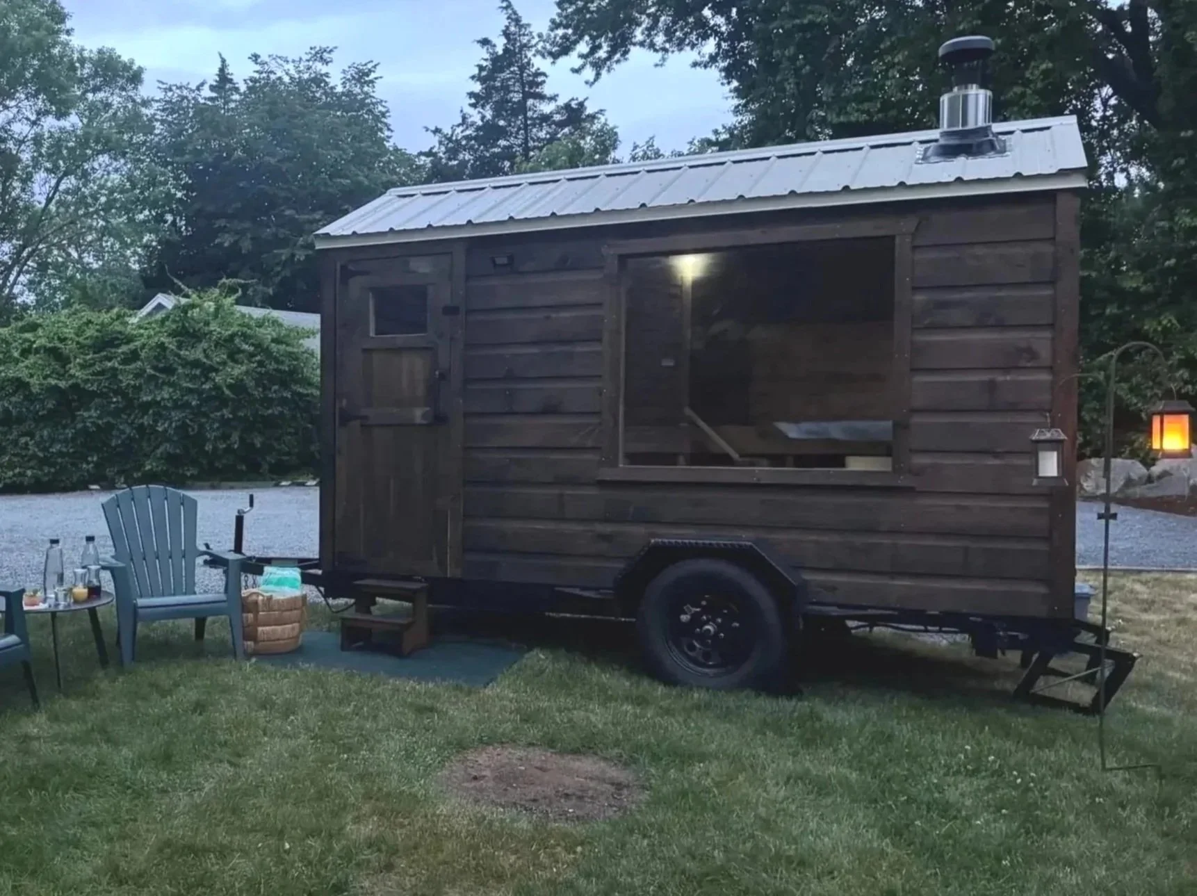 A small wooden food truck with a metal roof parked on a grassy area during dusk, surrounded by trees, with outdoor seating and decorative lighting.
