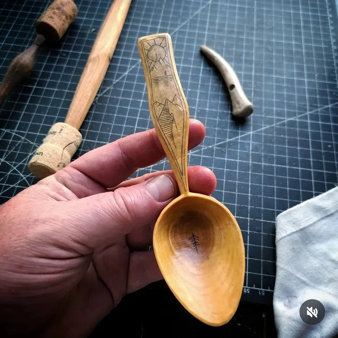 Hand holding a wooden spoon with carved mountain and sun design, surrounded by carving tools and a cloth on a grid work surface.