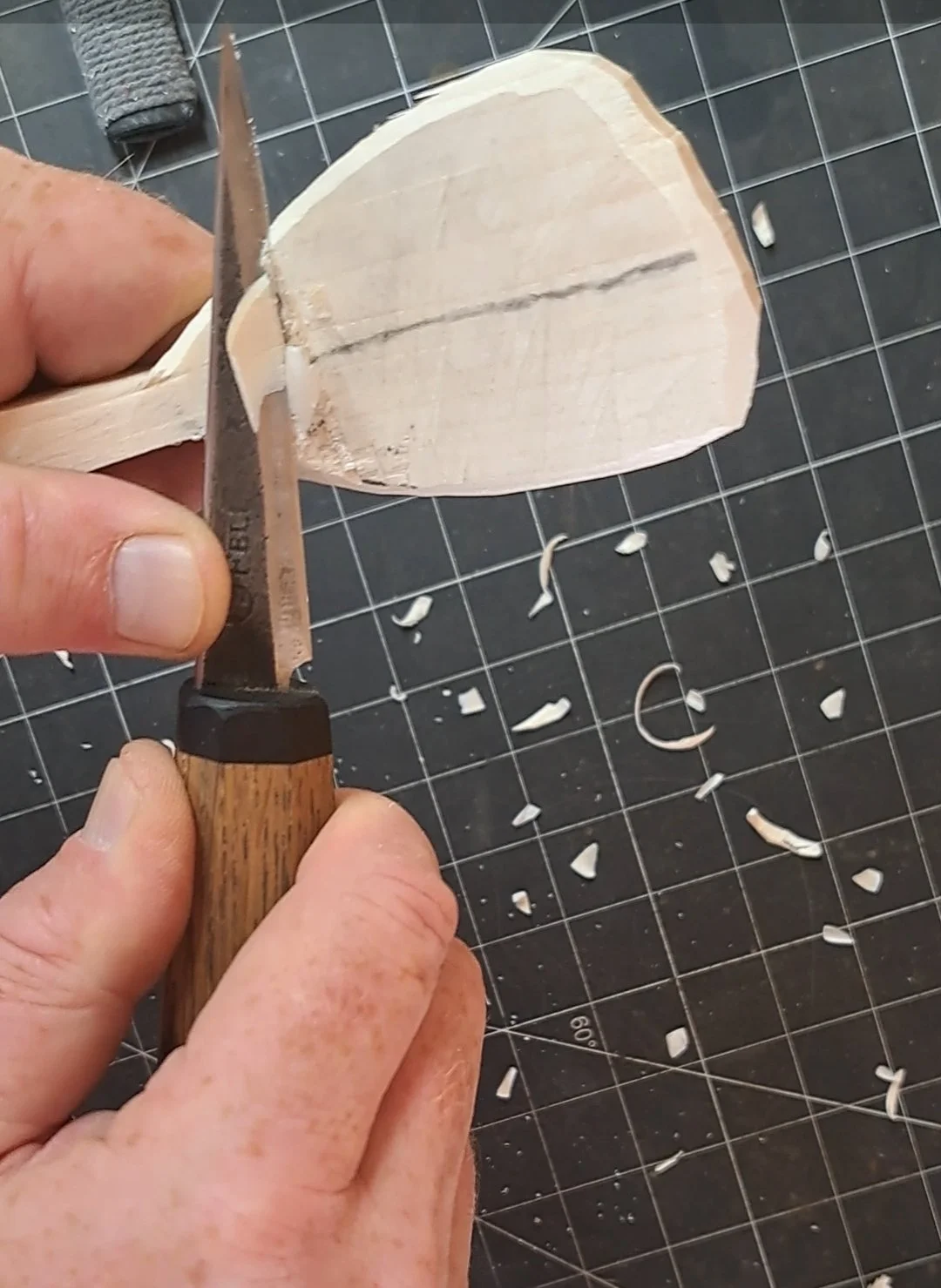 Close-up of a person's hands carving a piece of wood with a small straight knife, with wood shavings scattered on a cutting mat.