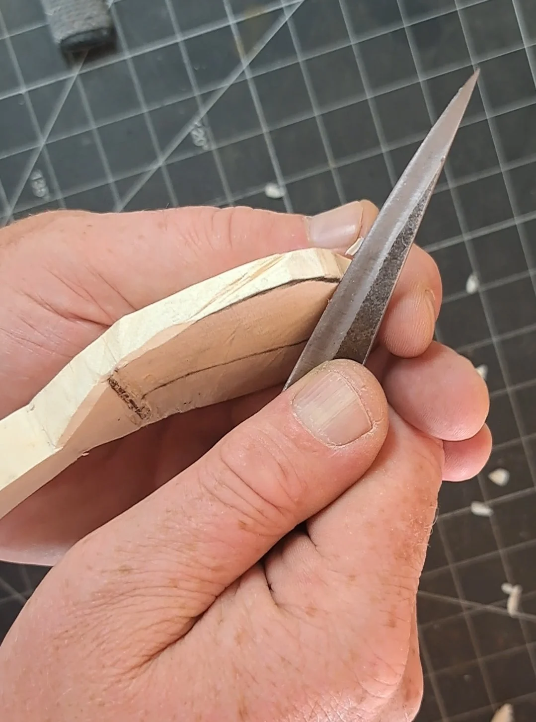 A craftsman carves the handle of a wooden object with a metal file, holding the piece firmly with his thumb and fingers. The background shows a black grid cutting mat.