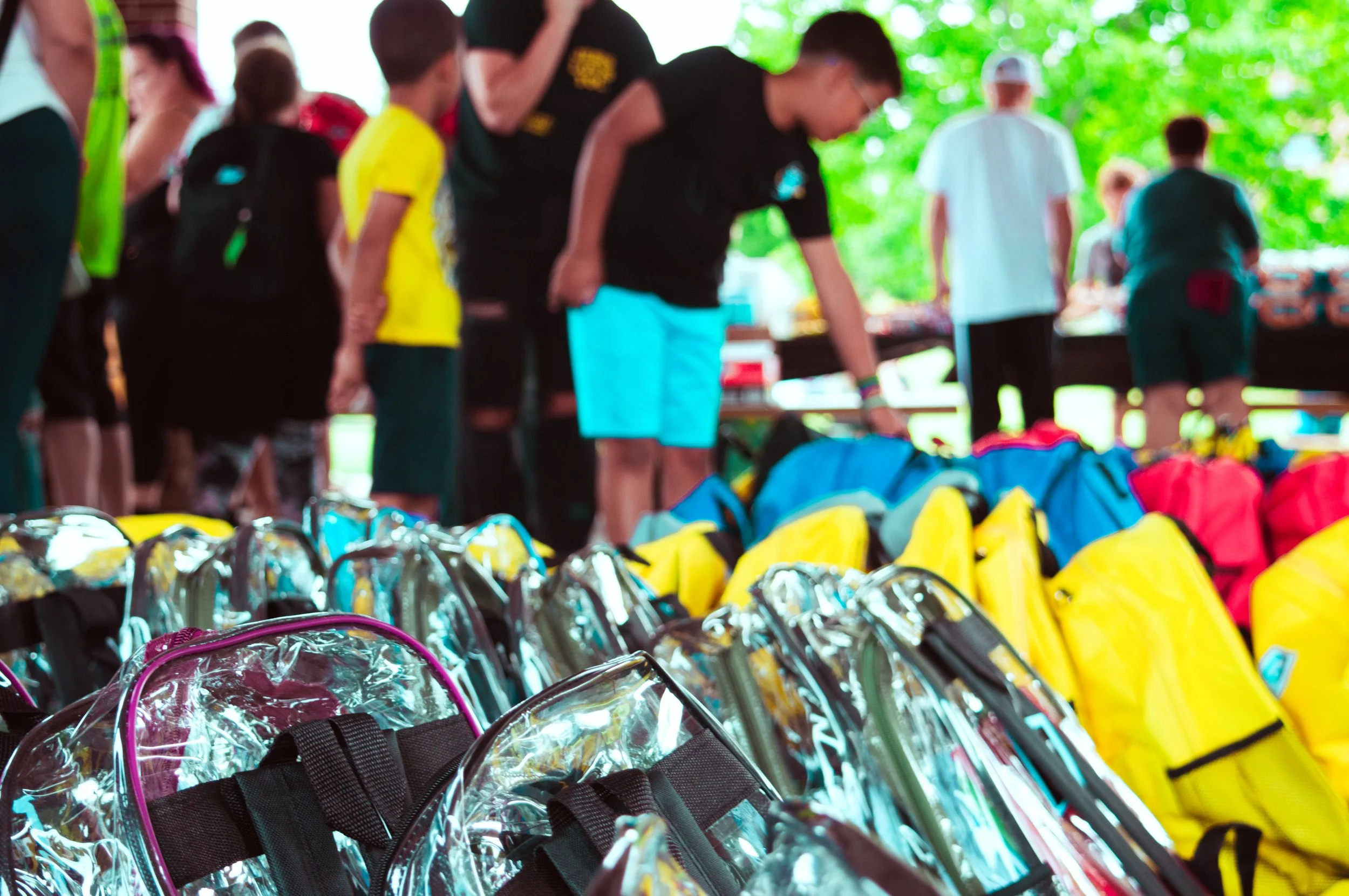 People browsing outdoor display of backpacks and gear at a fair or market, with blurred green trees in the background.