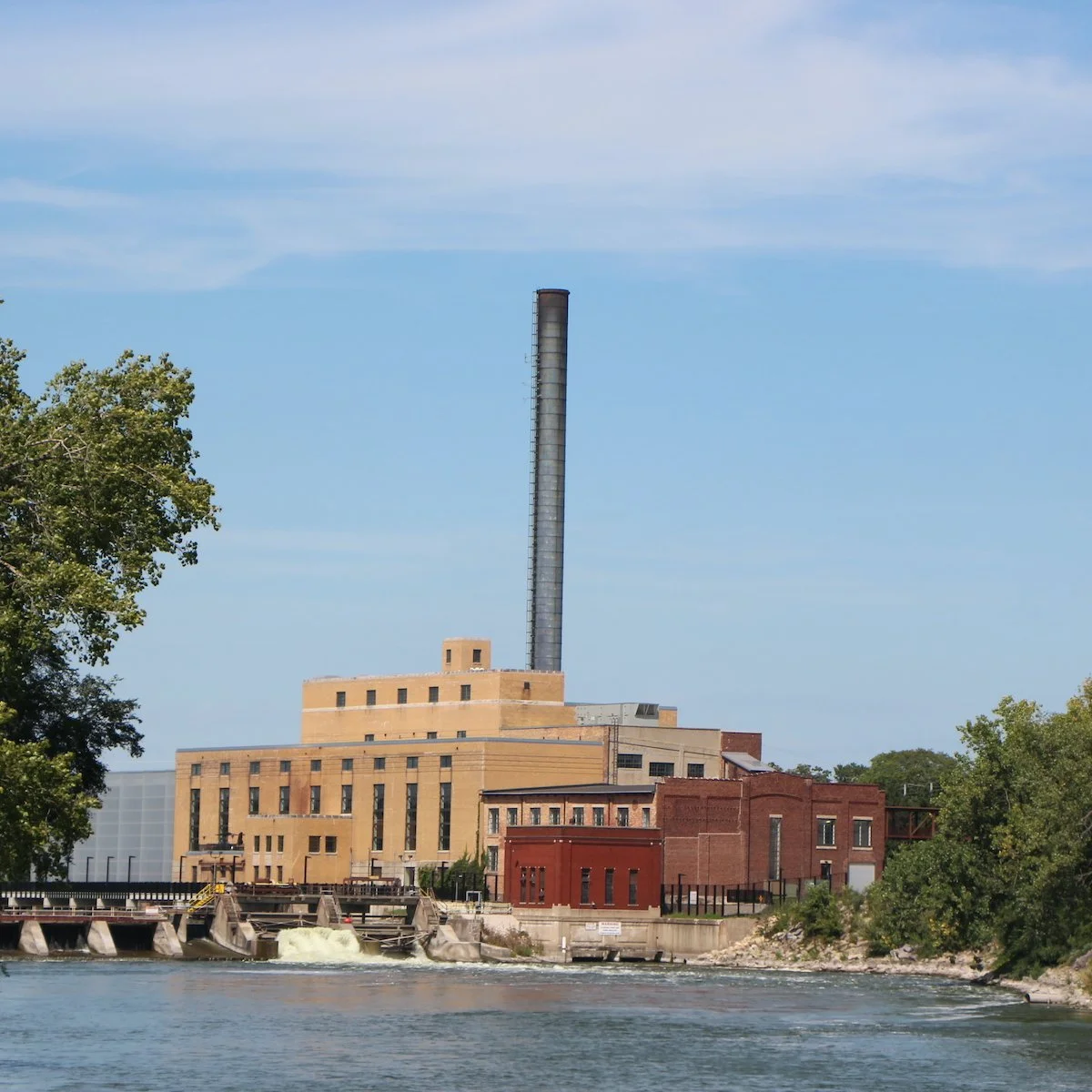 A historic industrial building with a tall chimney, situated near a river with trees on the sides, under a blue sky.