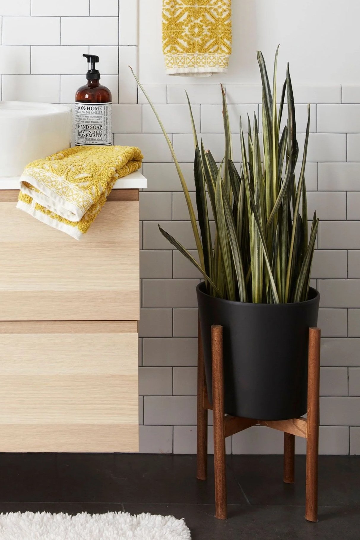 A bathroom with white subway tiles, a wooden vanity, and a black floor plant holder with a tall green plant. There is a yellow patterned towel hanging on the wall, a yellow hand towel folded on the vanity, and a bottle of hand soap.