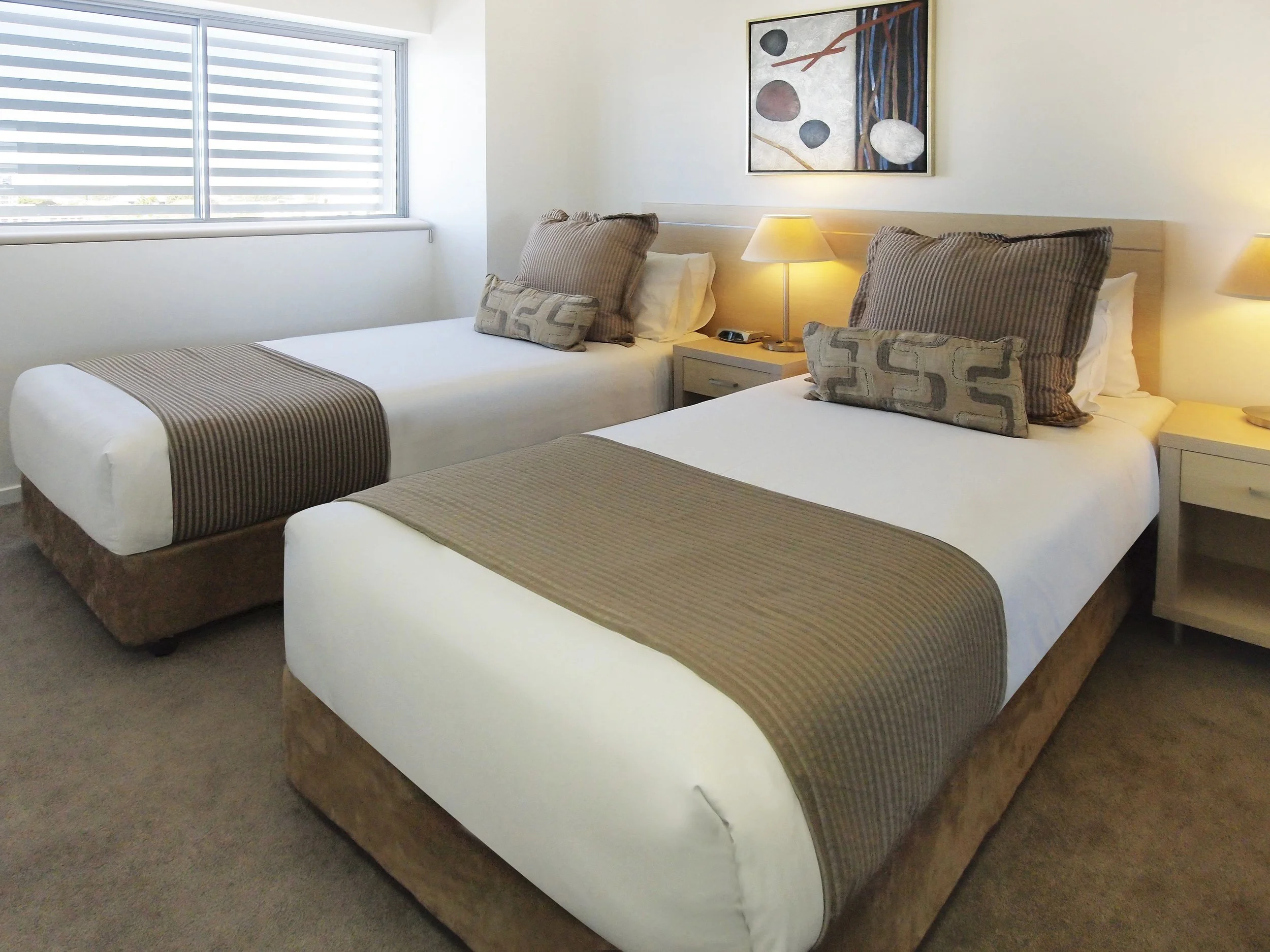 Two neatly made beds with beige and dark patterned pillows and beige bed runners in a hotel room, with a nightstand containing a lamp and an alarm clock between them, and a window with horizontal blinds on the wall.