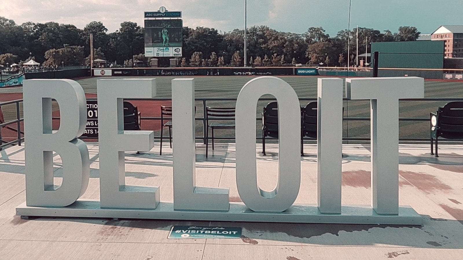 Large white sign spelling 'BELOIT' on a concrete platform at a baseball stadium, with a baseball field and scoreboard in the background.