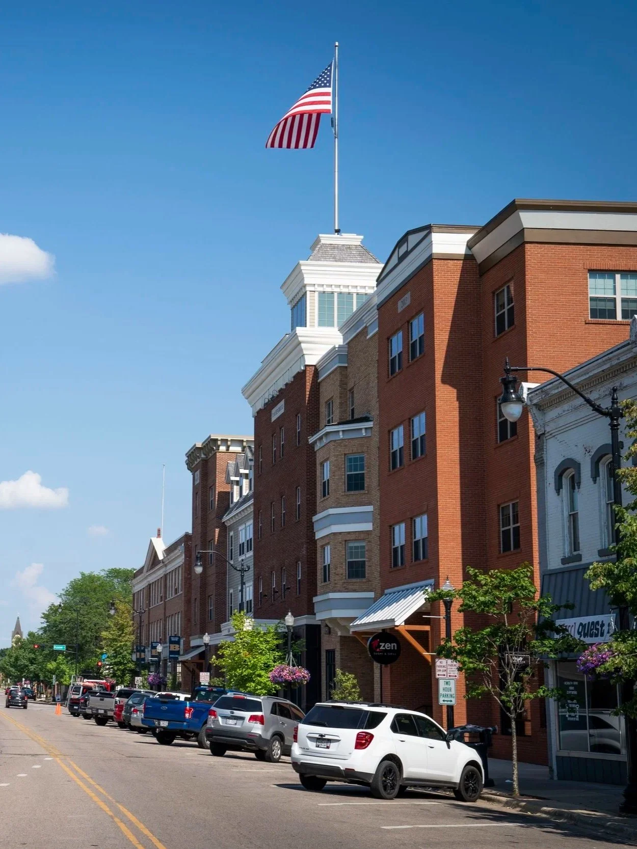 Street view of a downtown area with brick buildings, parked cars along the curb, and an American flag on a flagpole atop one of the buildings.