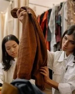 Two women shopping for clothes, one holding a brown jacket in a clothing store.