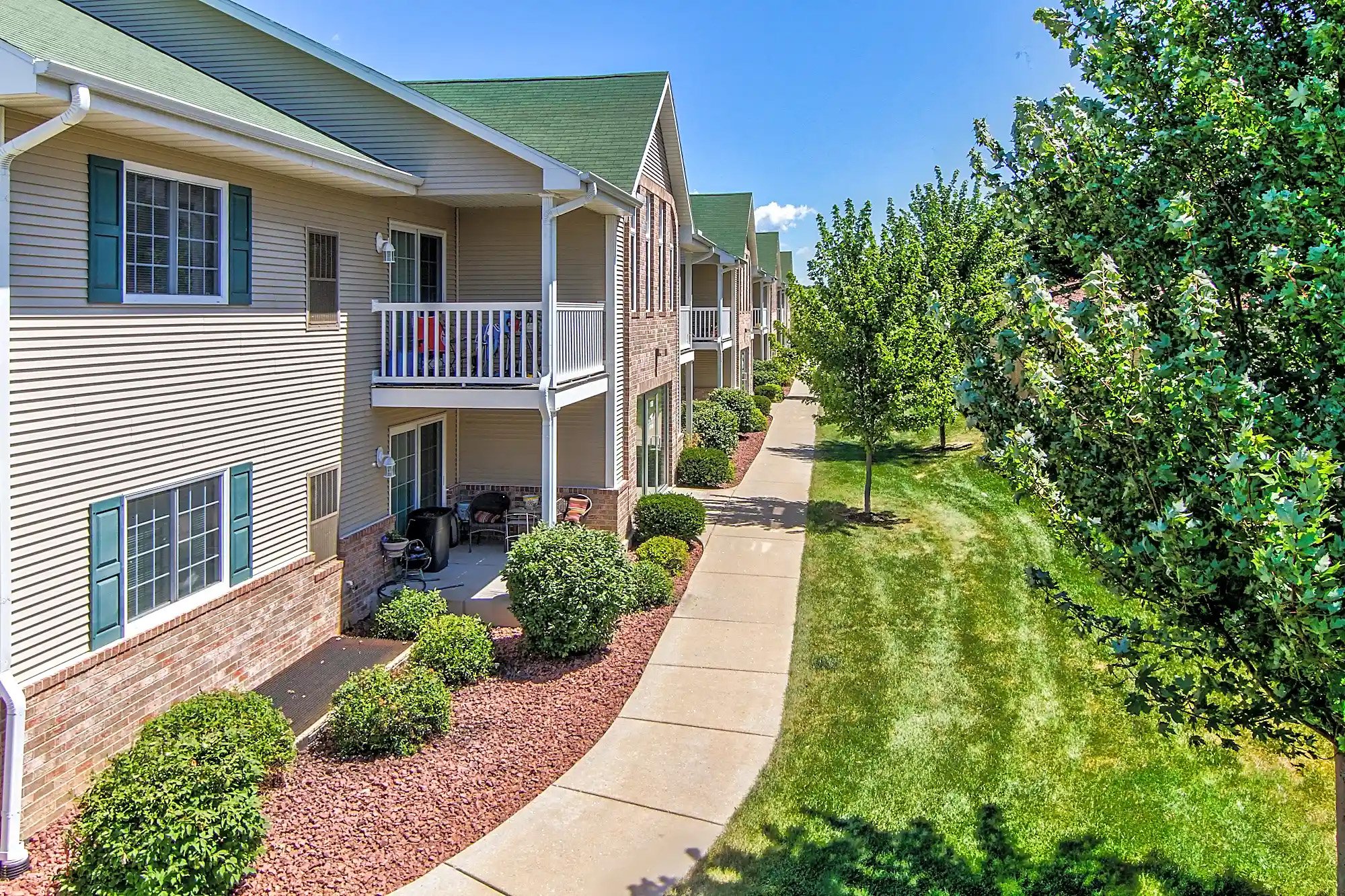 Exterior view of a townhouse complex with a sidewalk, green trees, well-kept lawn, and a bright blue sky.