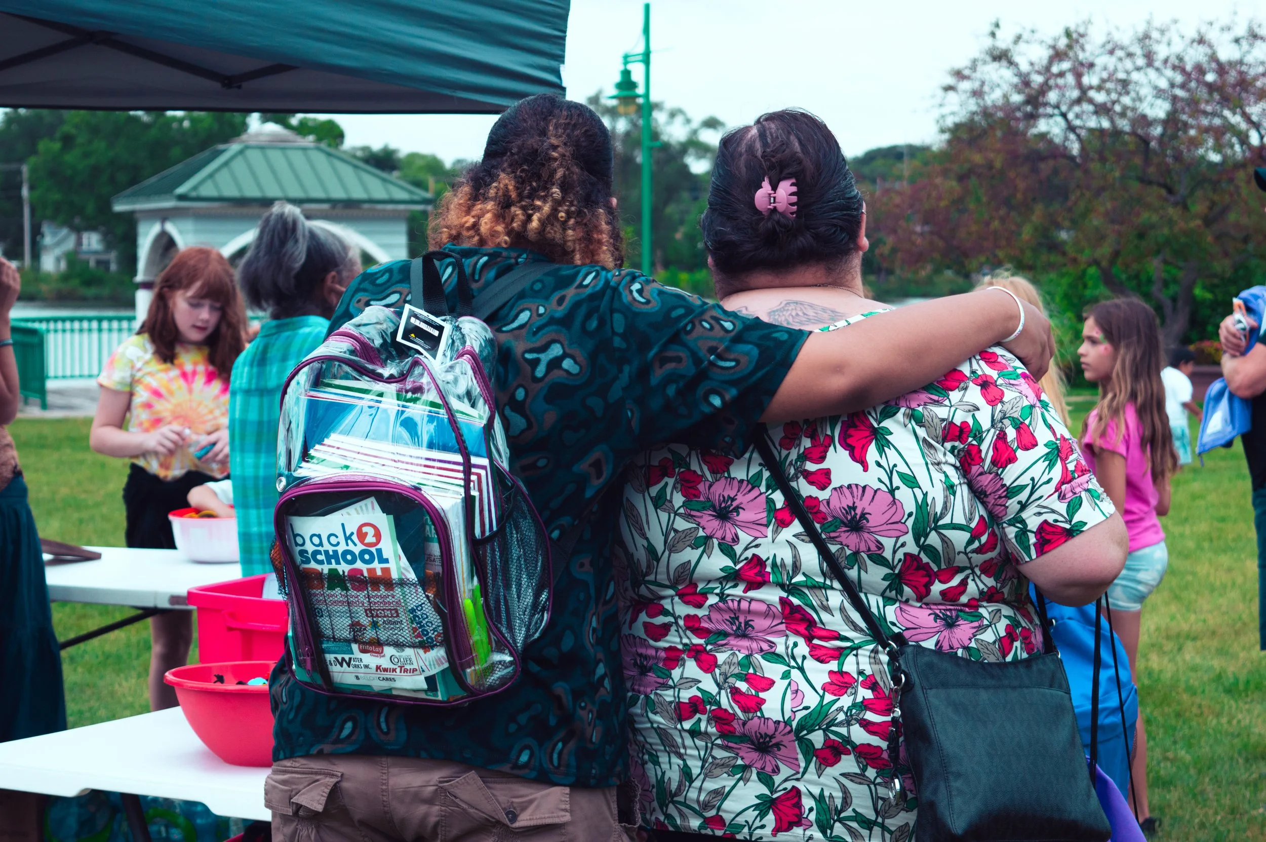 People gathered outdoors around a table with backpacks, some children and adults embracing at a back-to-school event with trees and a gazebo in the background.