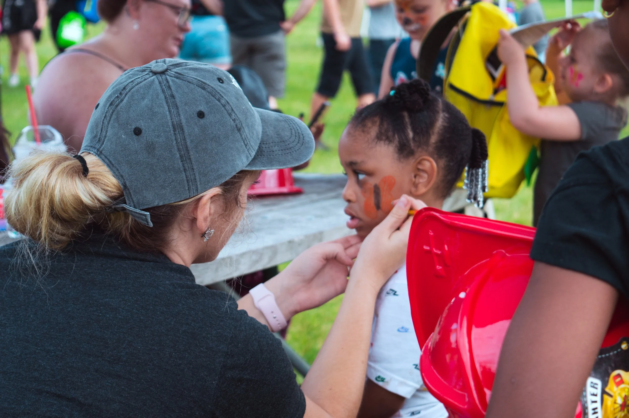 A woman with a gray cap is face painting a young girl with a white shirt and a red chair, at an outdoor event. The girl has orange and black face paint on her cheek and is sitting still as the woman applies more face paint. Several other children and adults are visible in the background.