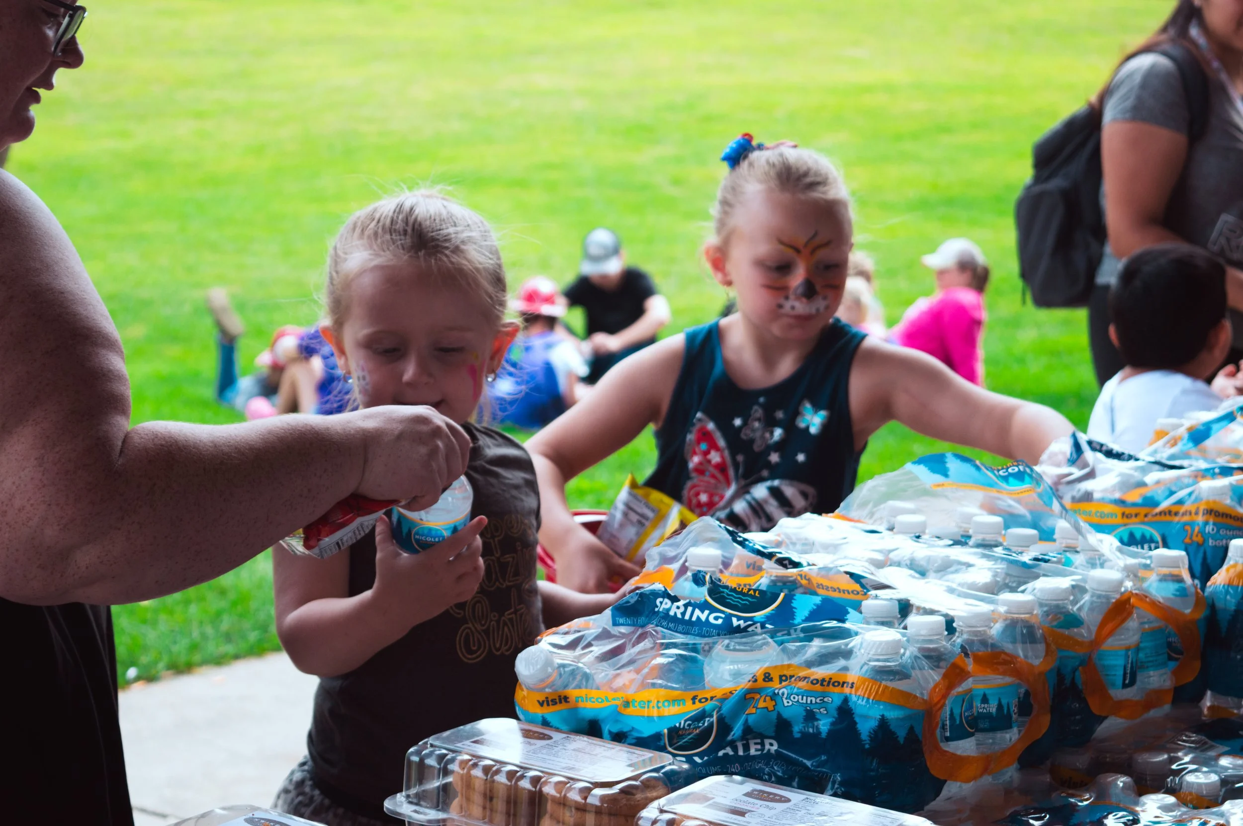 Children and adults gather around a table outdoors, distributing bottled water and snacks, with face paint on some children, on a grassy area during daytime.