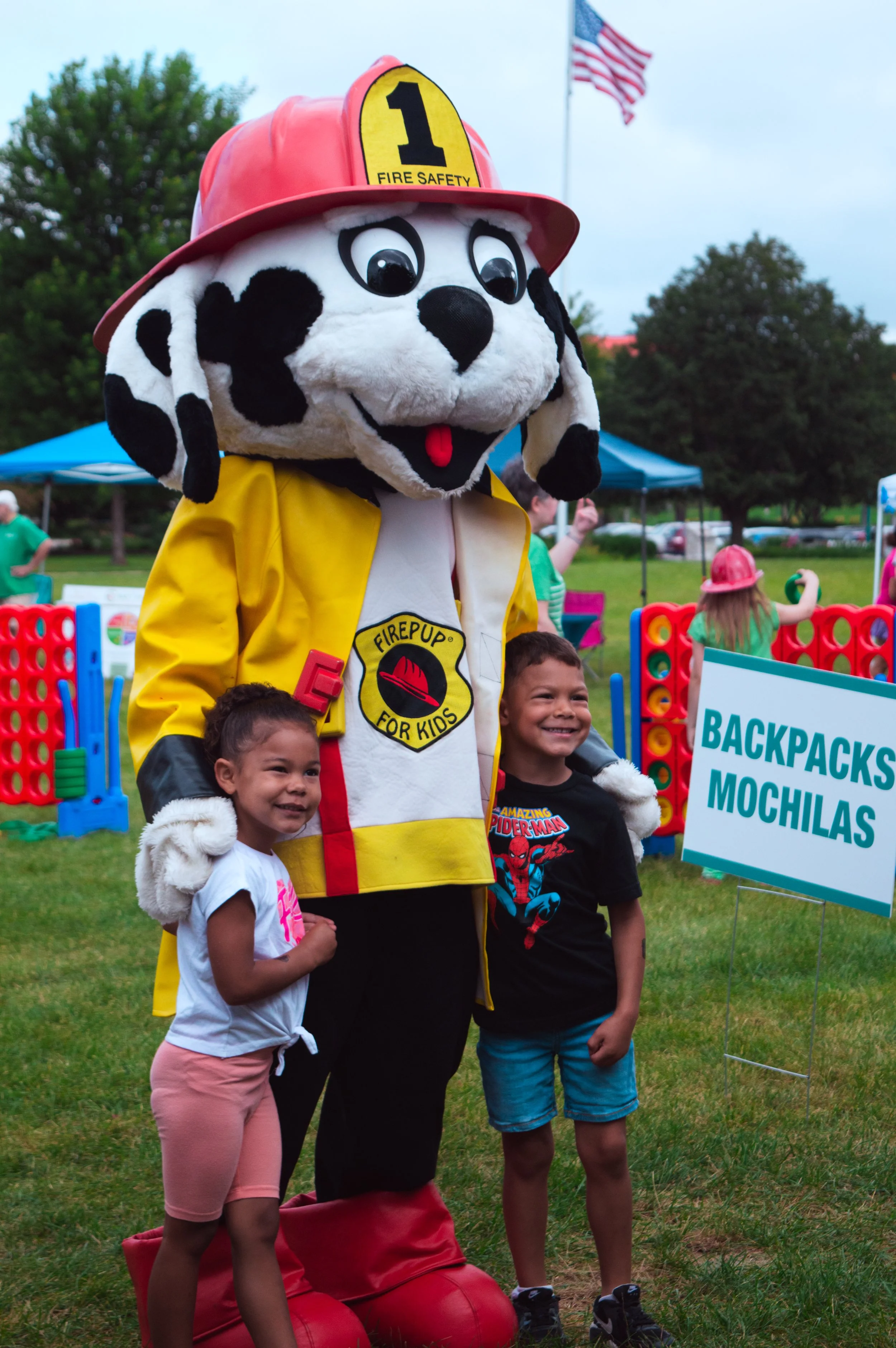 Children with a rescue mascot at an outdoor event, mascot wearing a firefighter helmet and yellow shirt, children smiling and posing.