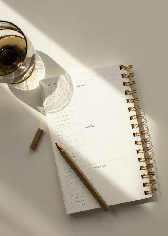 Open planner with days listed from Monday to Wednesday, a gold pen, and a glass of tea or coffee on a white surface with sunlight.
