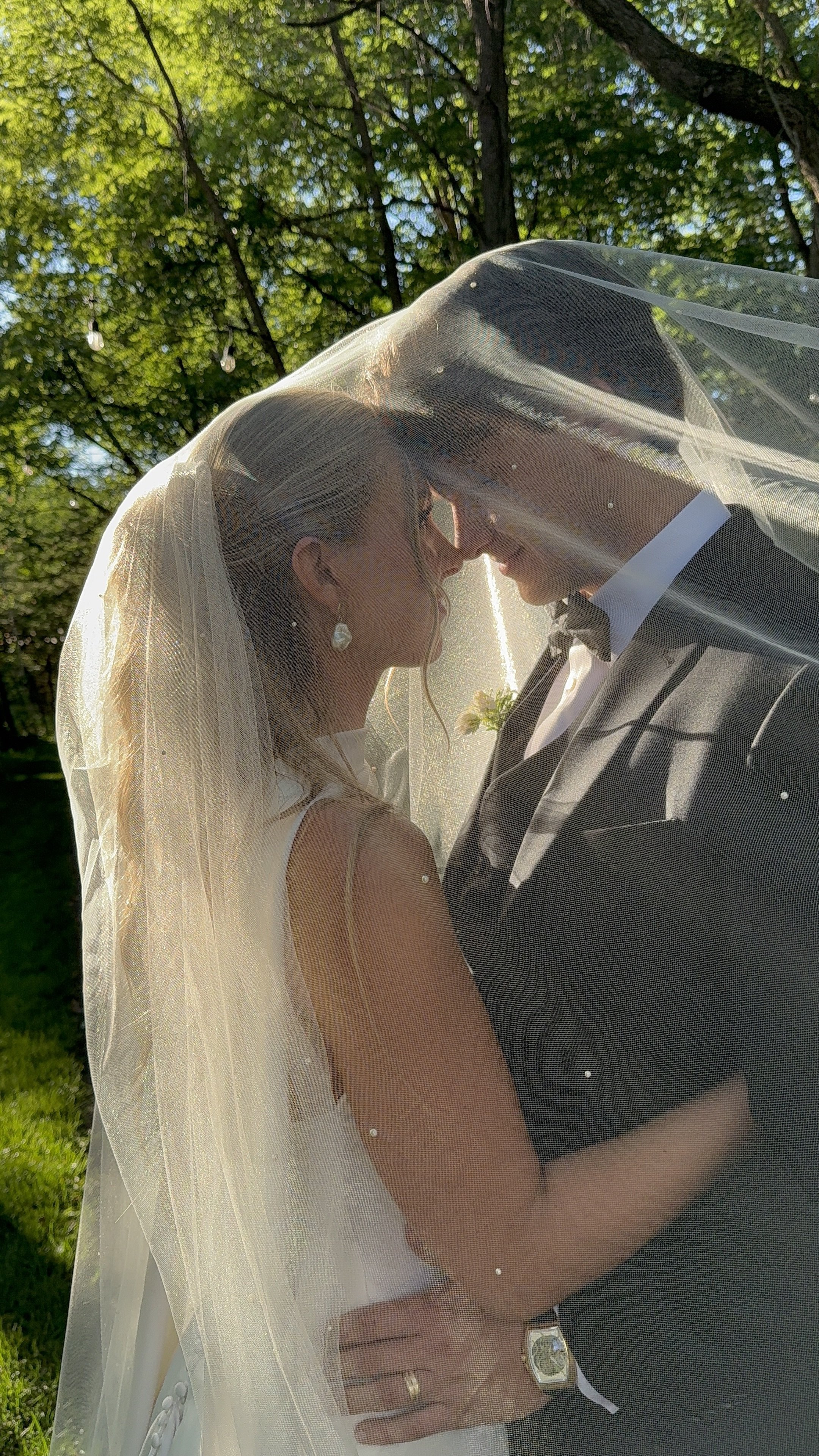 A bride and groom standing face-to-face outdoors under a canopy of green leaves, sharing an intimate moment with their foreheads touching, seen through a sheer veil.