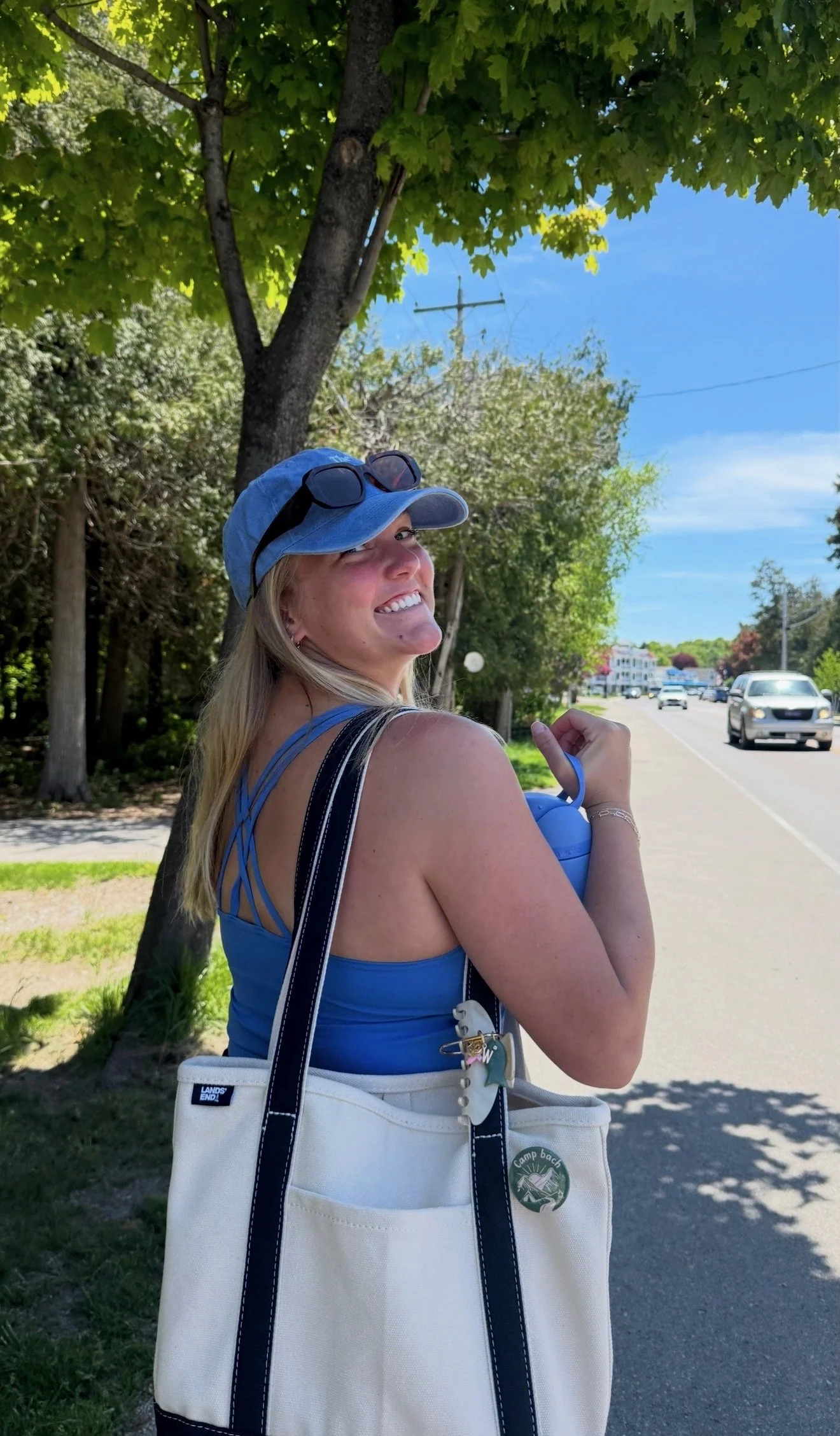 Smiling woman wearing a blue cap with sunglasses on top, blue tank top, and white bag with a camp badge, standing outdoors on a sunny day next to a tree and a street with cars.