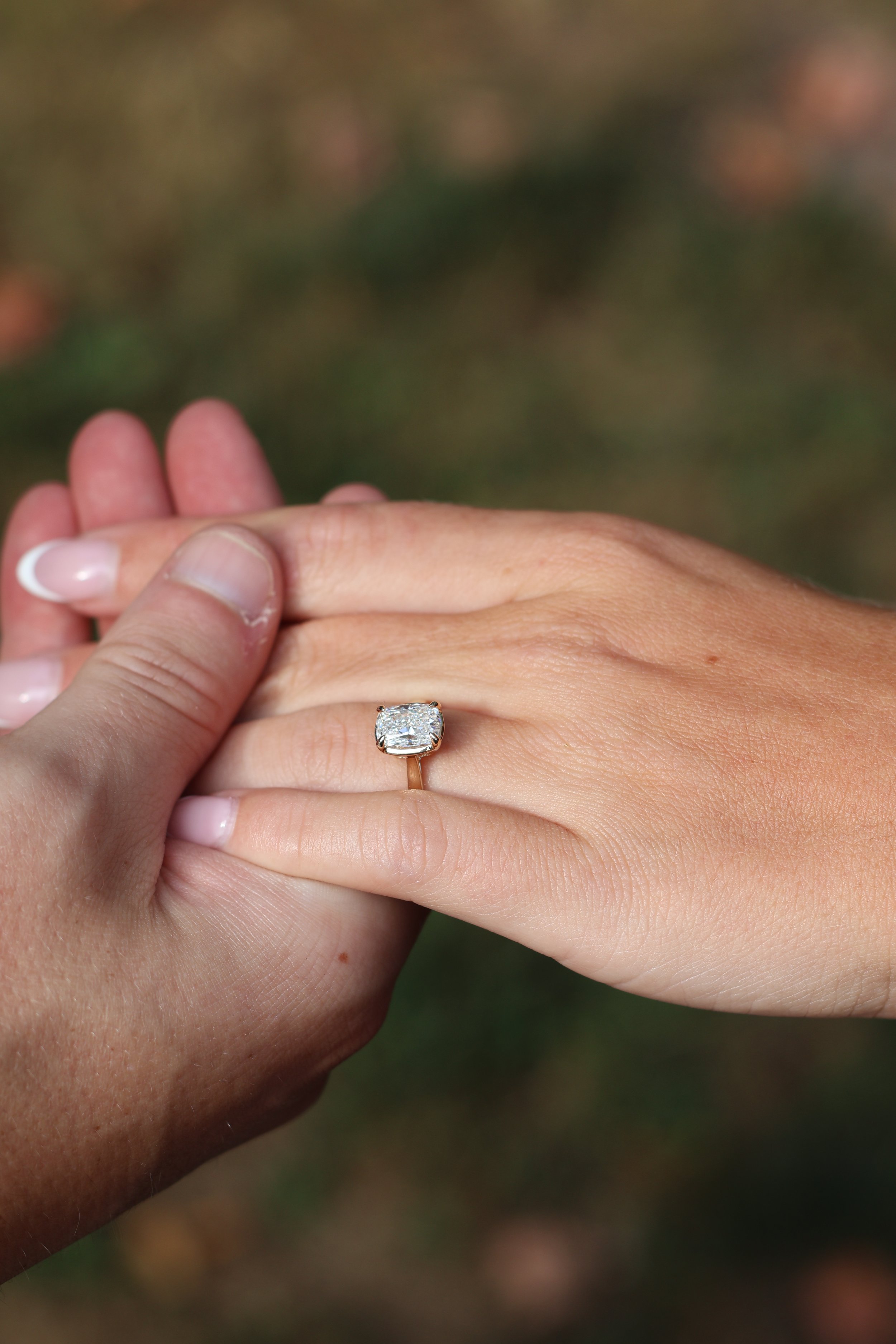 Close-up of two hands gently holding each other, with a focus on a ring with a large square-cut diamond on the finger of one hand.