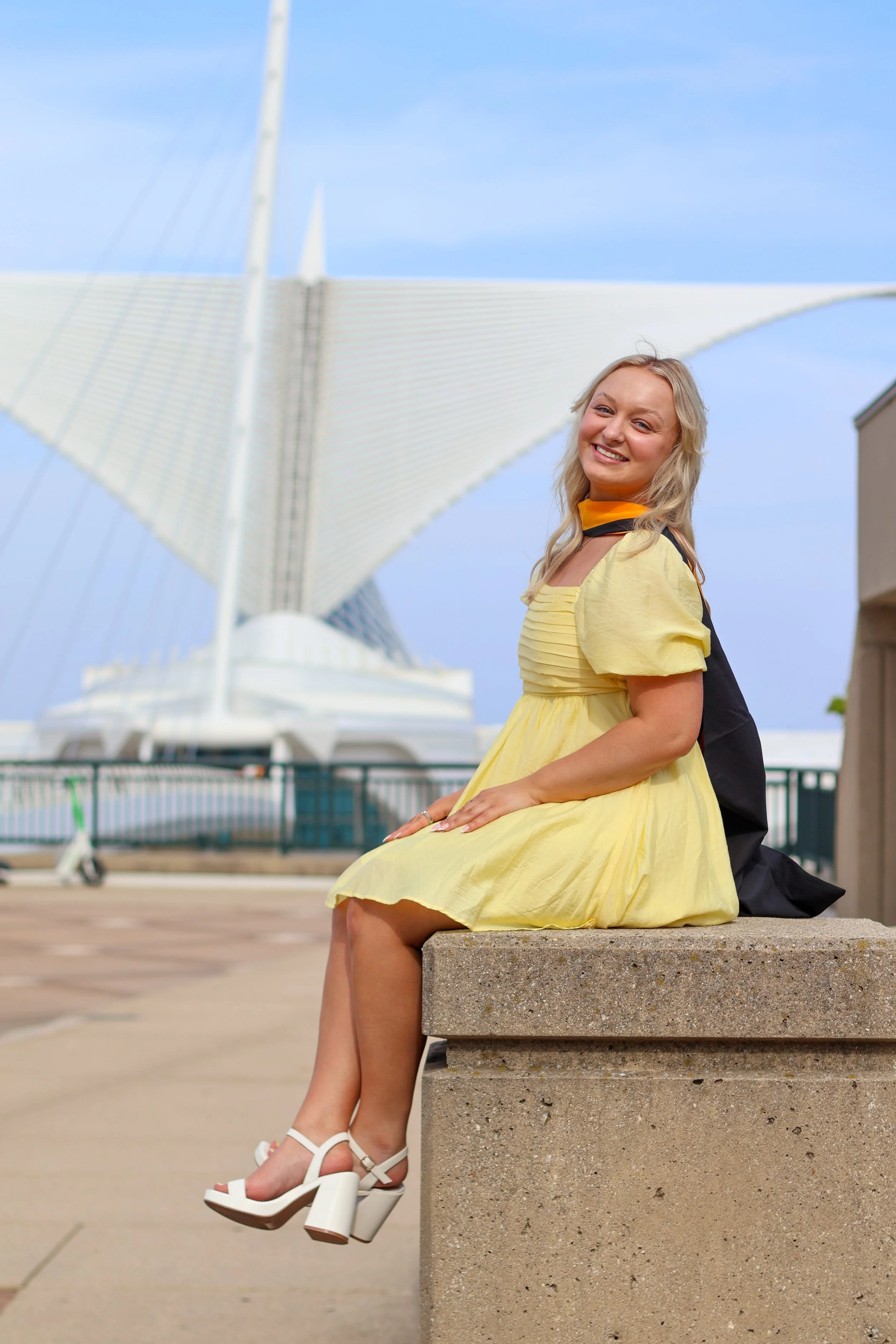 A woman in a yellow dress with white high heels sitting on a concrete bench by the water with a modern bridge in the background.