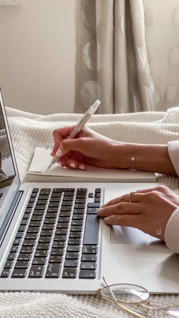 A person working on a laptop, writing in a notebook with a pen, with glasses resting nearby on a textured blanket, and beige curtains in the background.