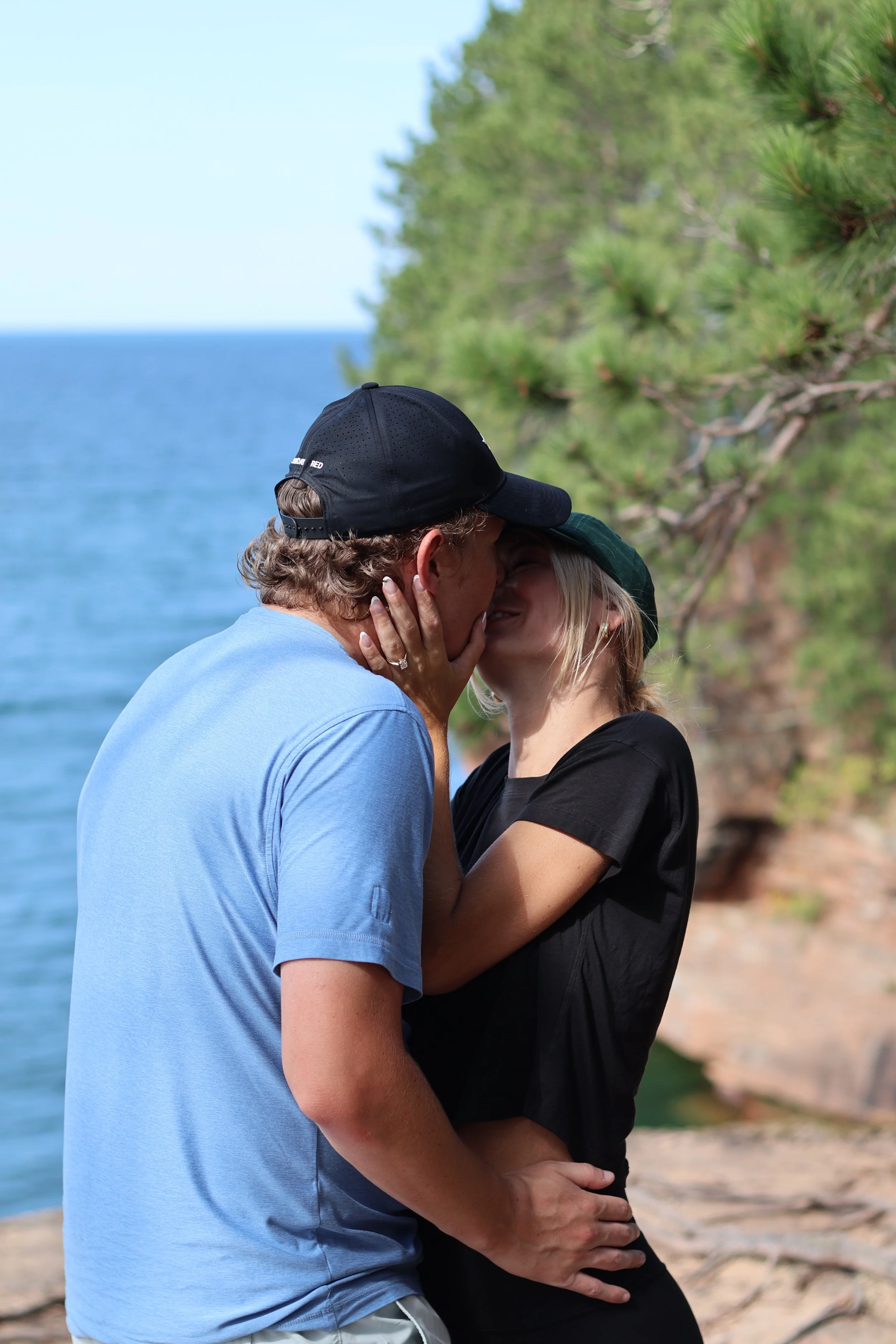 A couple kissing on a beach with trees and the ocean in the background.