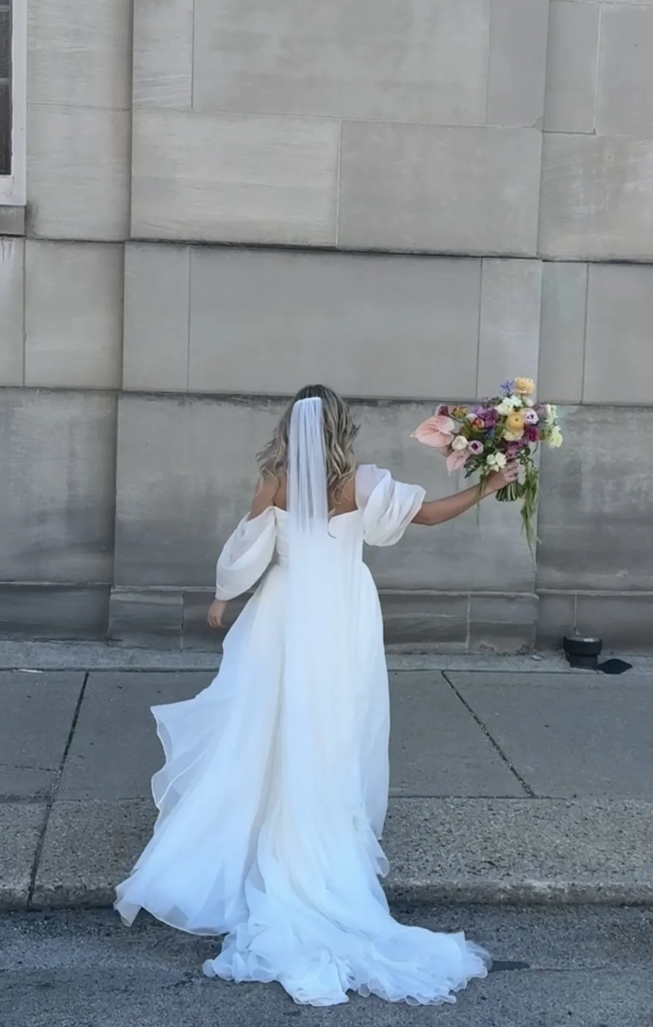 A woman in a white wedding dress holding a bouquet of mixed flowers stands with her back to the camera in front of a stone wall.