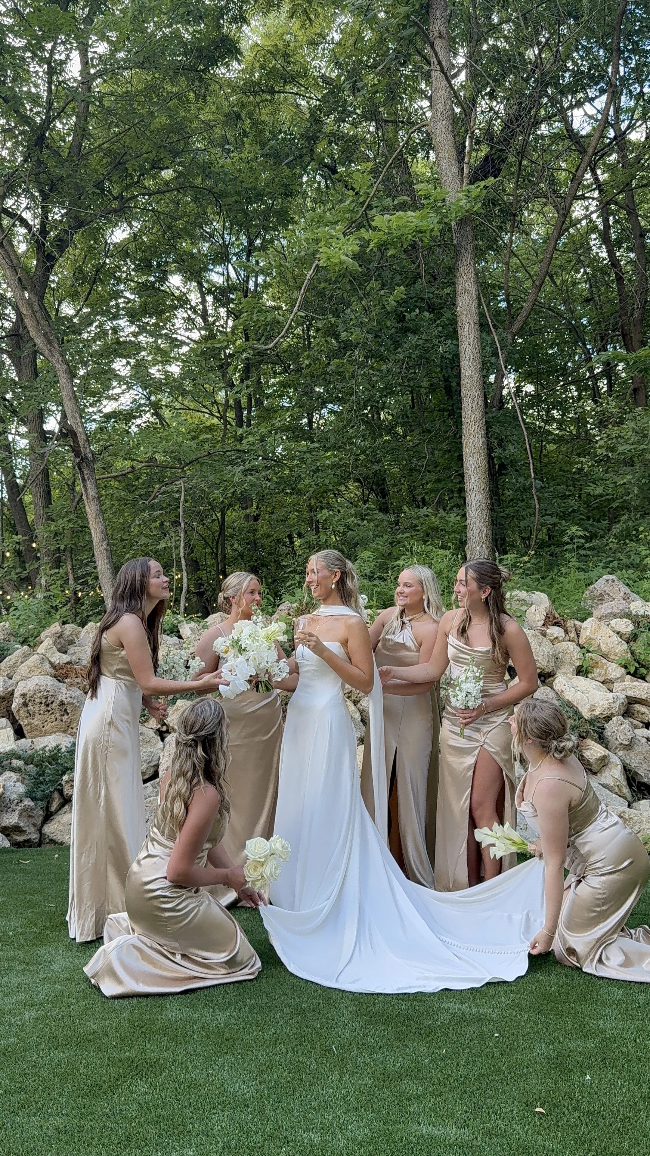 A group of women at an outdoor wedding ceremony, with one bride in a white wedding dress and the others in matching beige dresses, holding bouquets of white flowers, surrounded by rocks and trees.