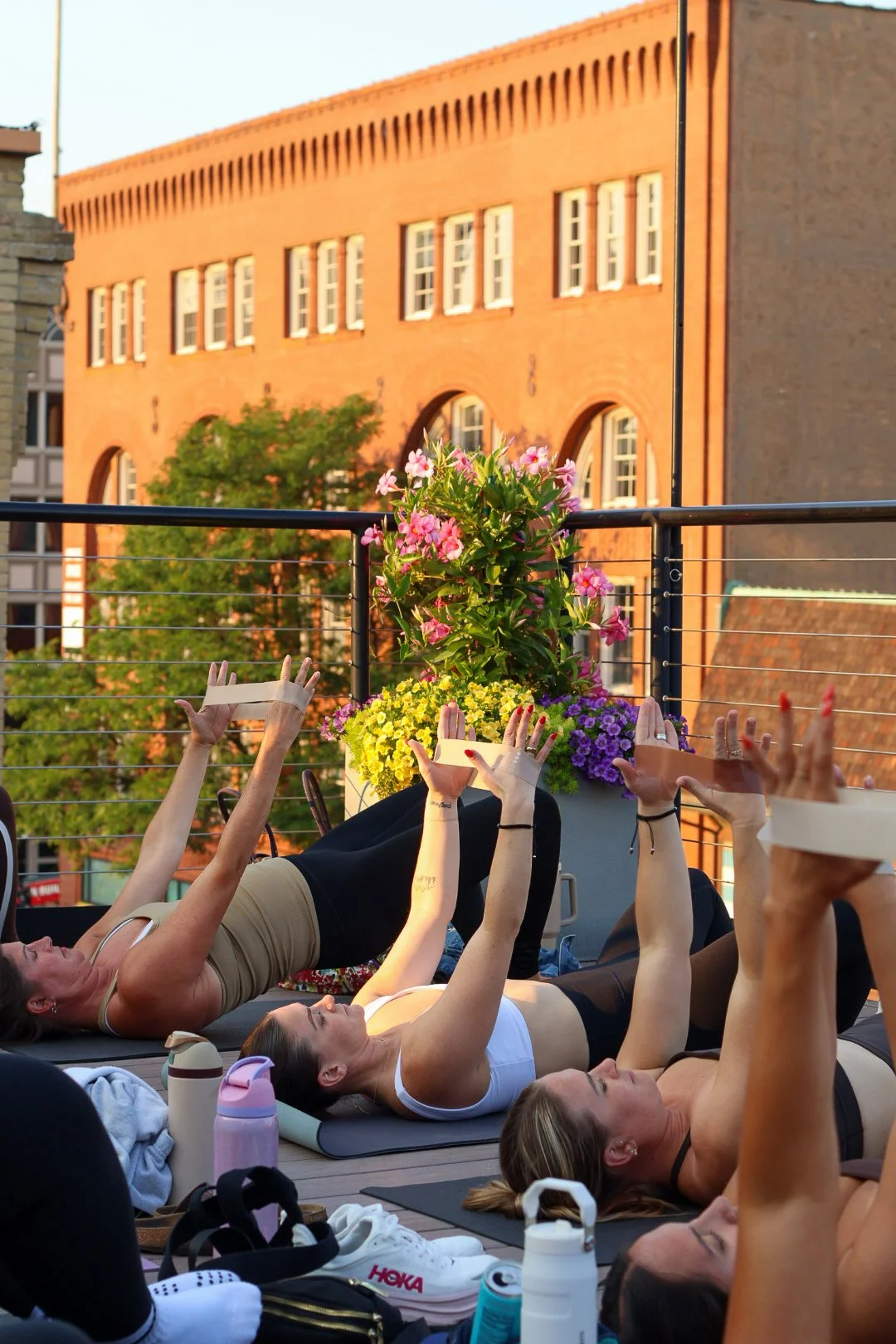People practicing yoga or stretching on a rooftop during sunset, with a large flowerspot and city buildings in the background.