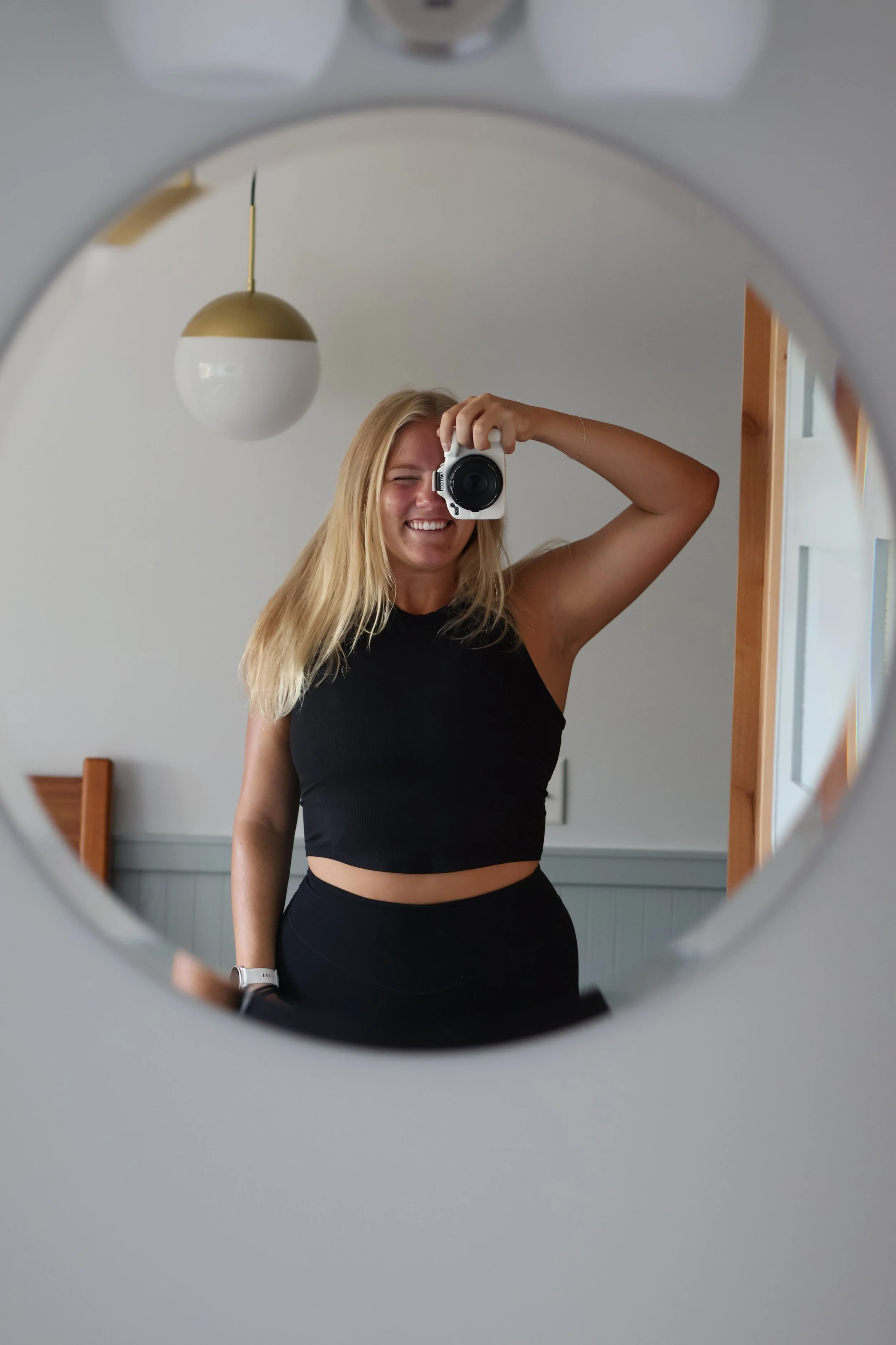 A young woman takes a mirror selfie with a camera, smiling while wearing a black sleeveless crop top and black pants, in a room with white walls and a wooden door frame.